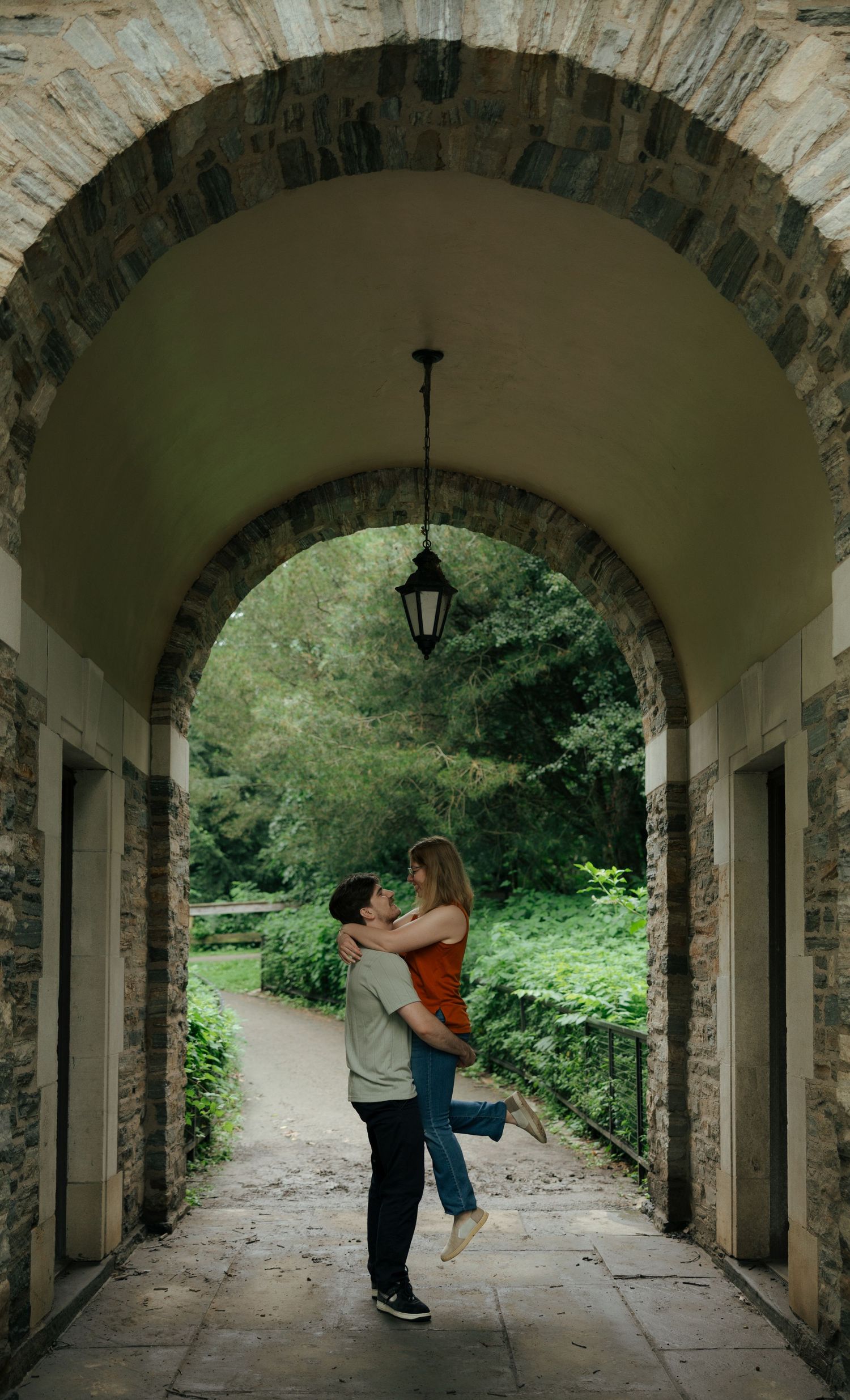 A couple shares a romantic lift and kiss under a stone archway surrounded by lush greenery.