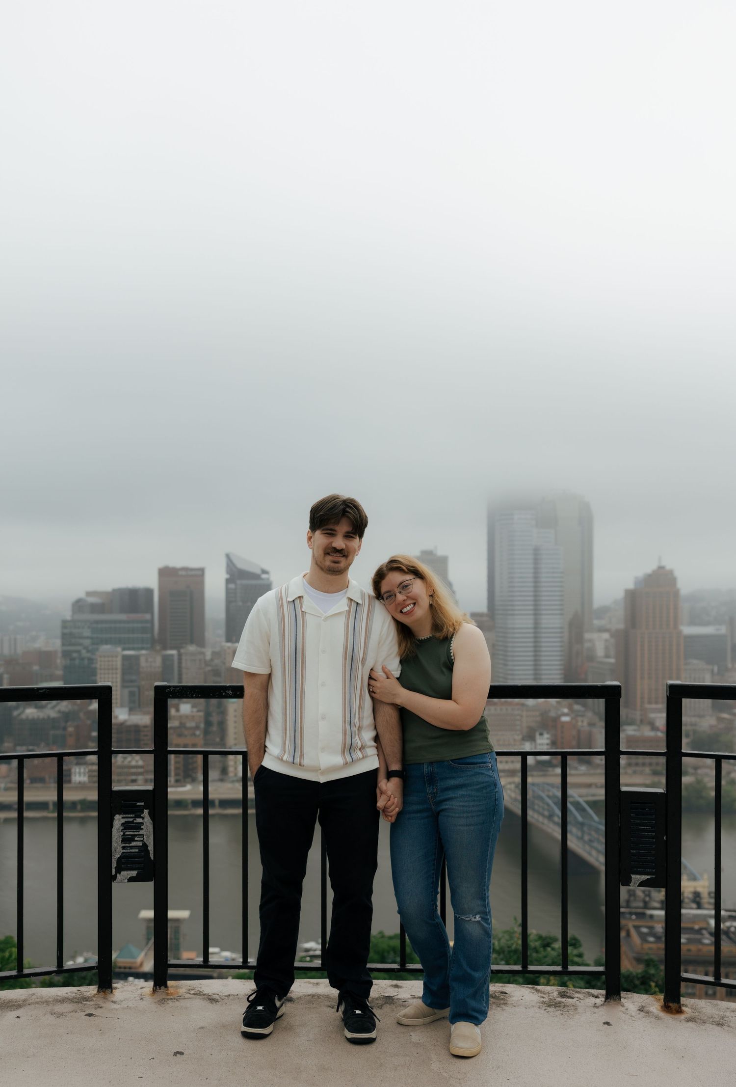 Couple posing on a scenic overlook with a misty city skyline and river view behind them.