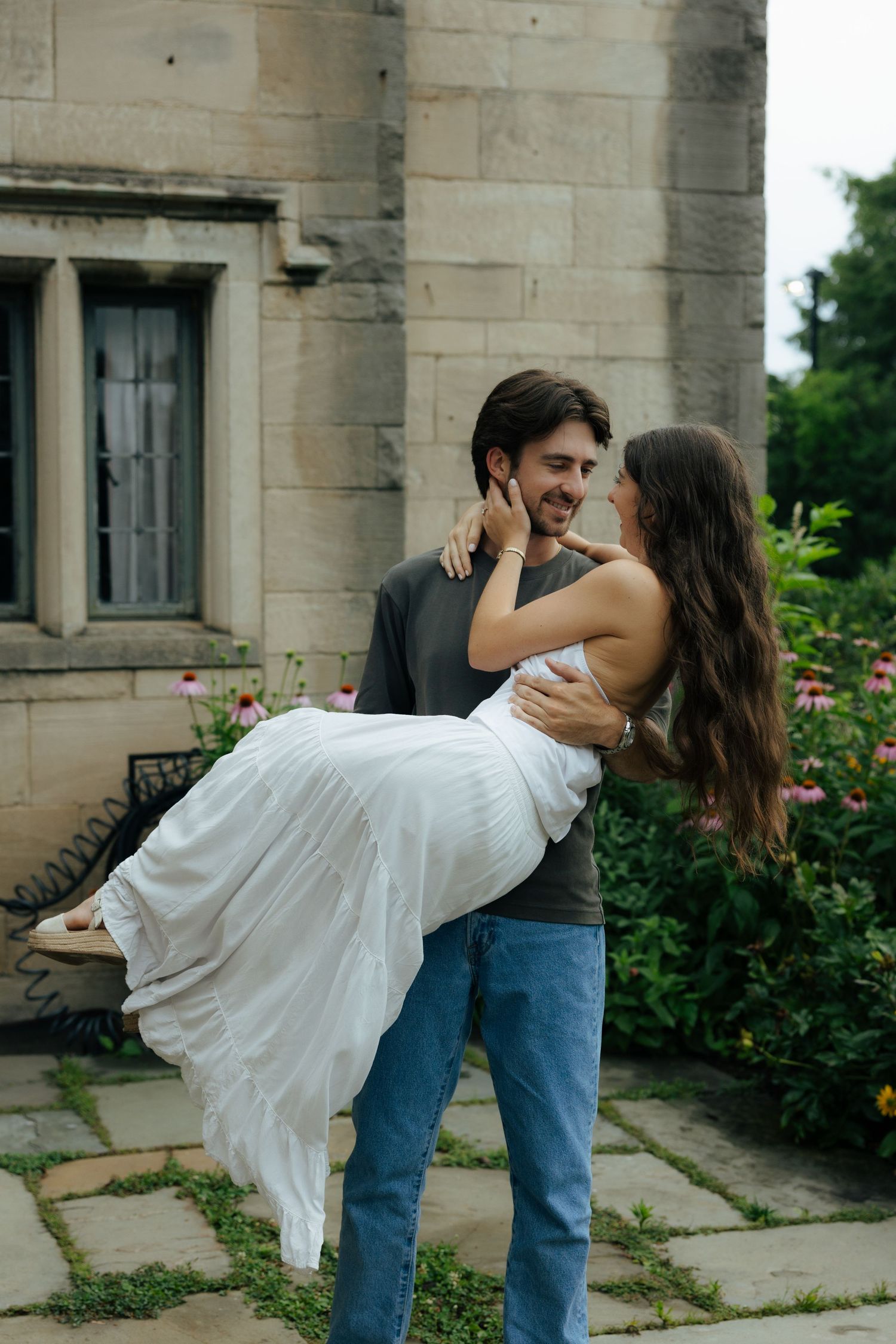 A man lifts a laughing woman in a white dress outside a stone building surrounded by garden flowers.