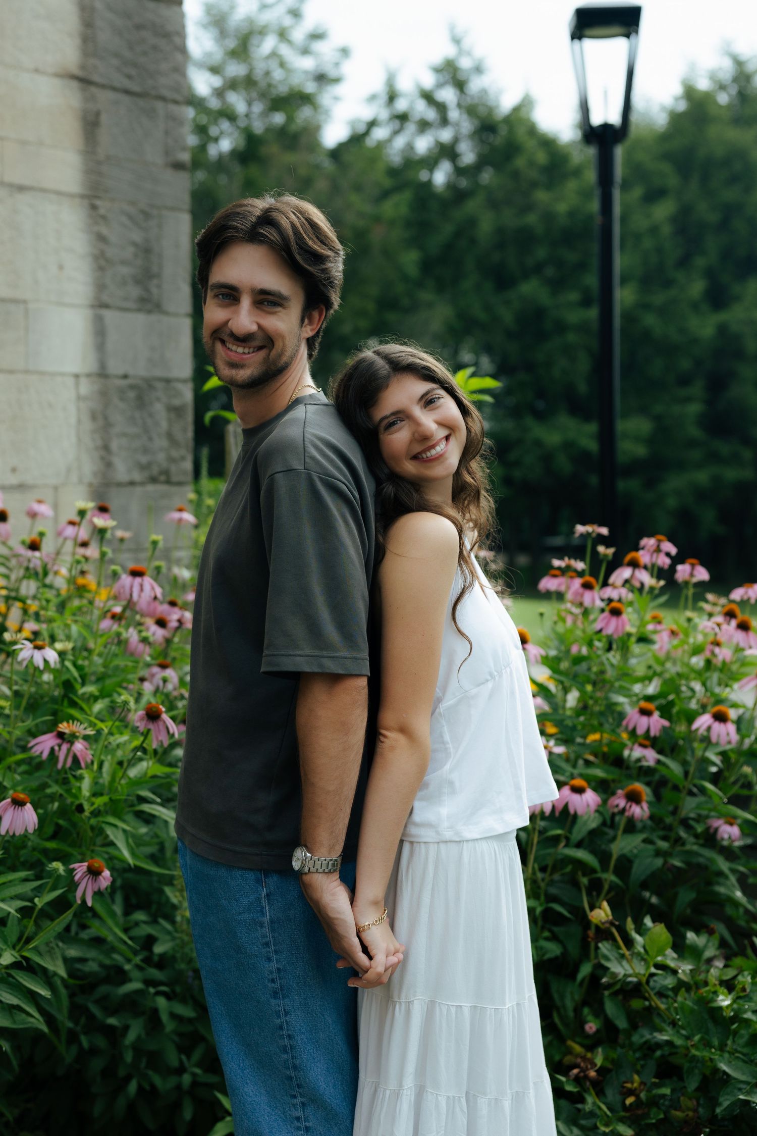 A smiling couple holds hands while standing among pink coneflowers in a garden.