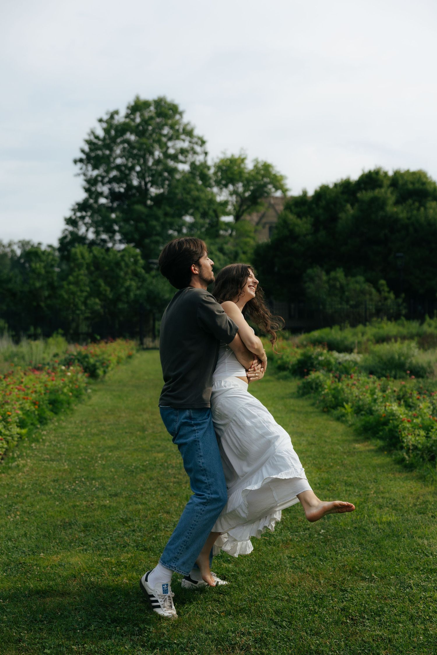Couple dancing and spinning joyfully on a green garden path lined with flowering plants.