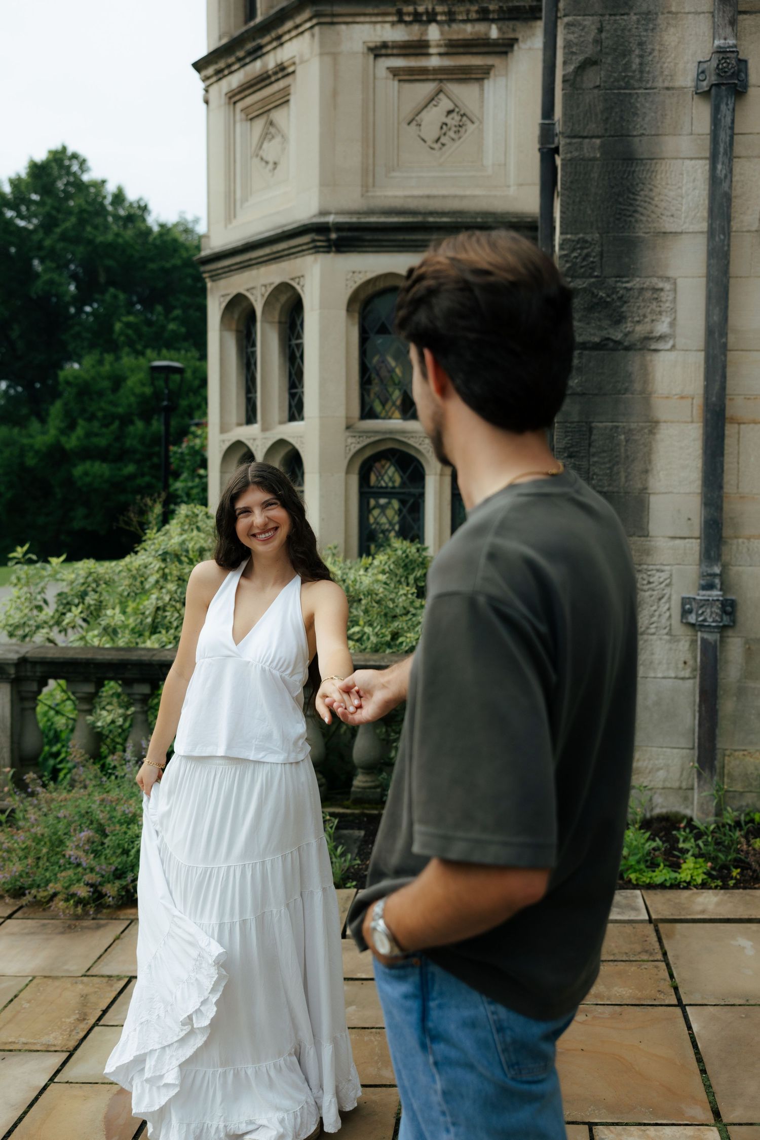 Smiling woman in white dress holds hands with man in gray tee outside a historic stone building.