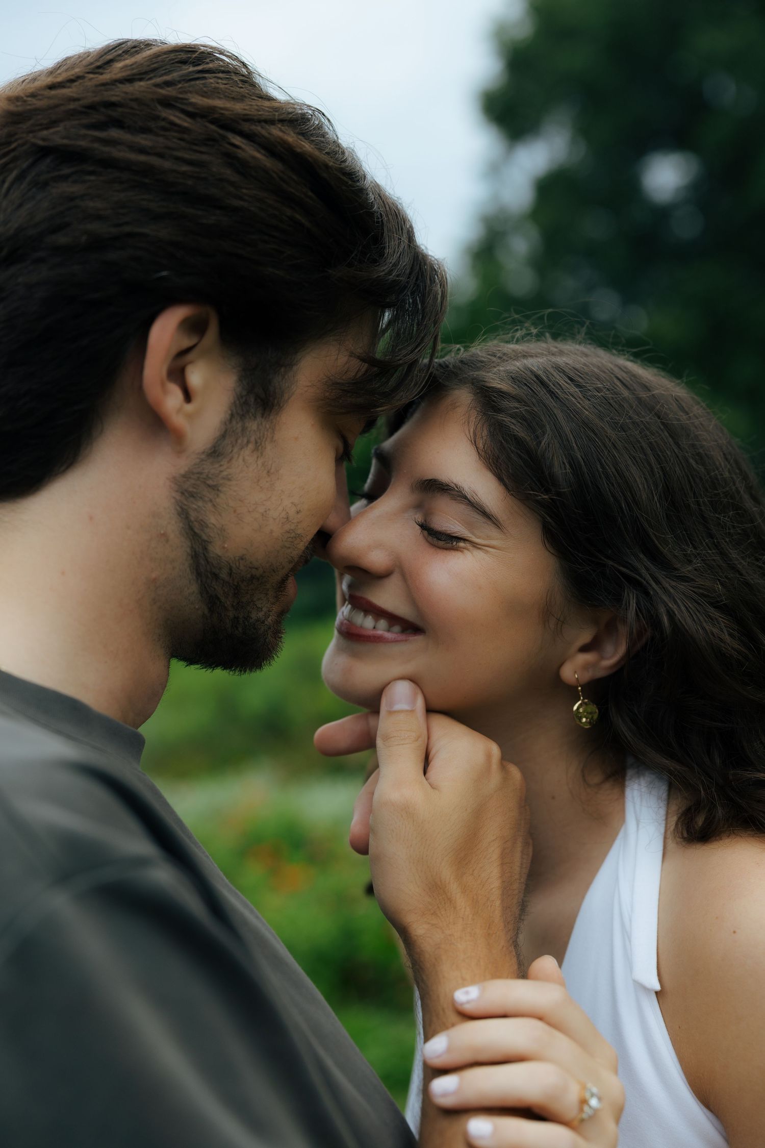 Romantic couple nearly kissing outdoors, smiling woman with gold earring and engagement ring held by bearded man.