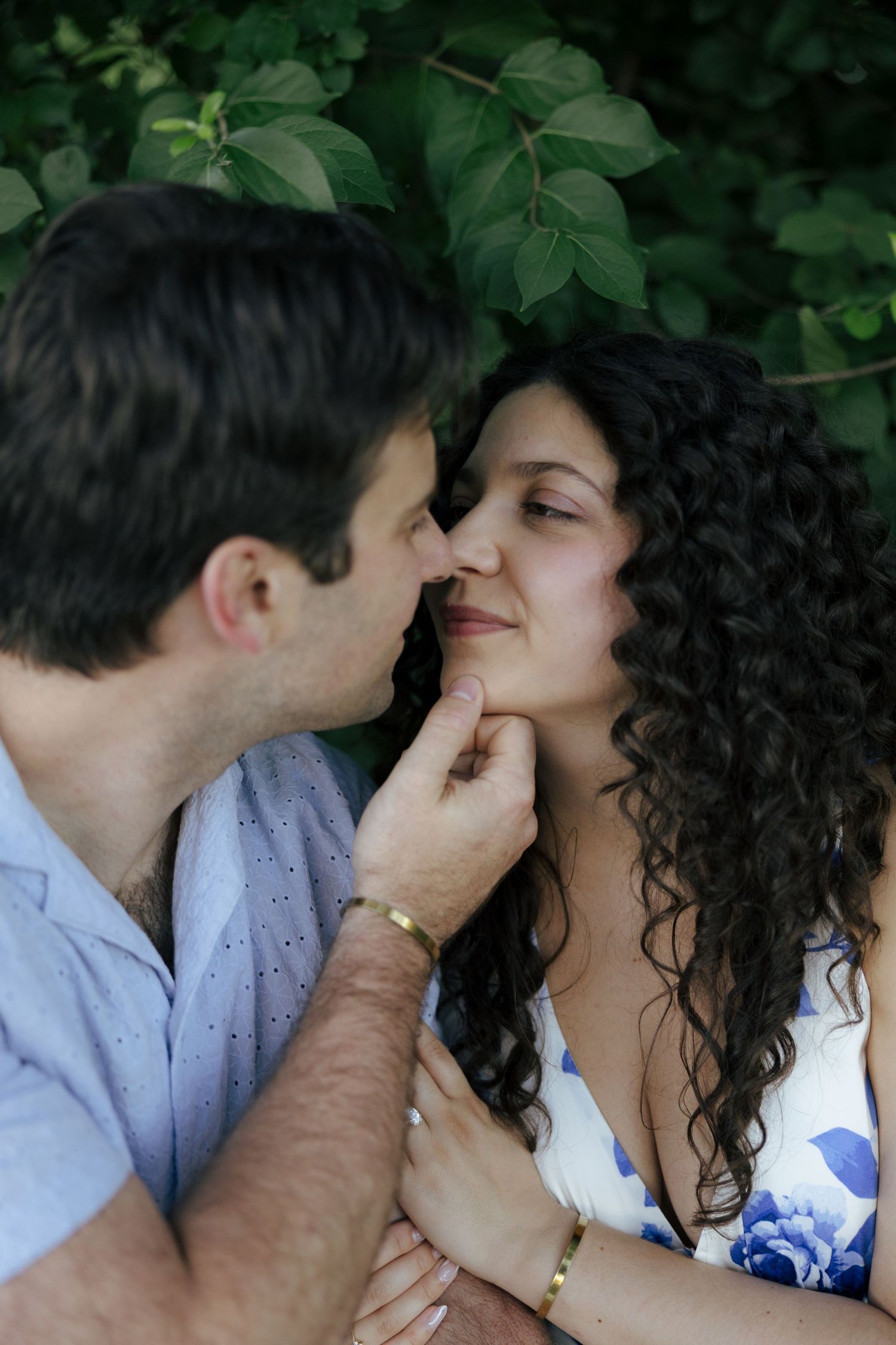 A couple shares a tender moment outdoors, man gently touching womans chin near lush green foliage.