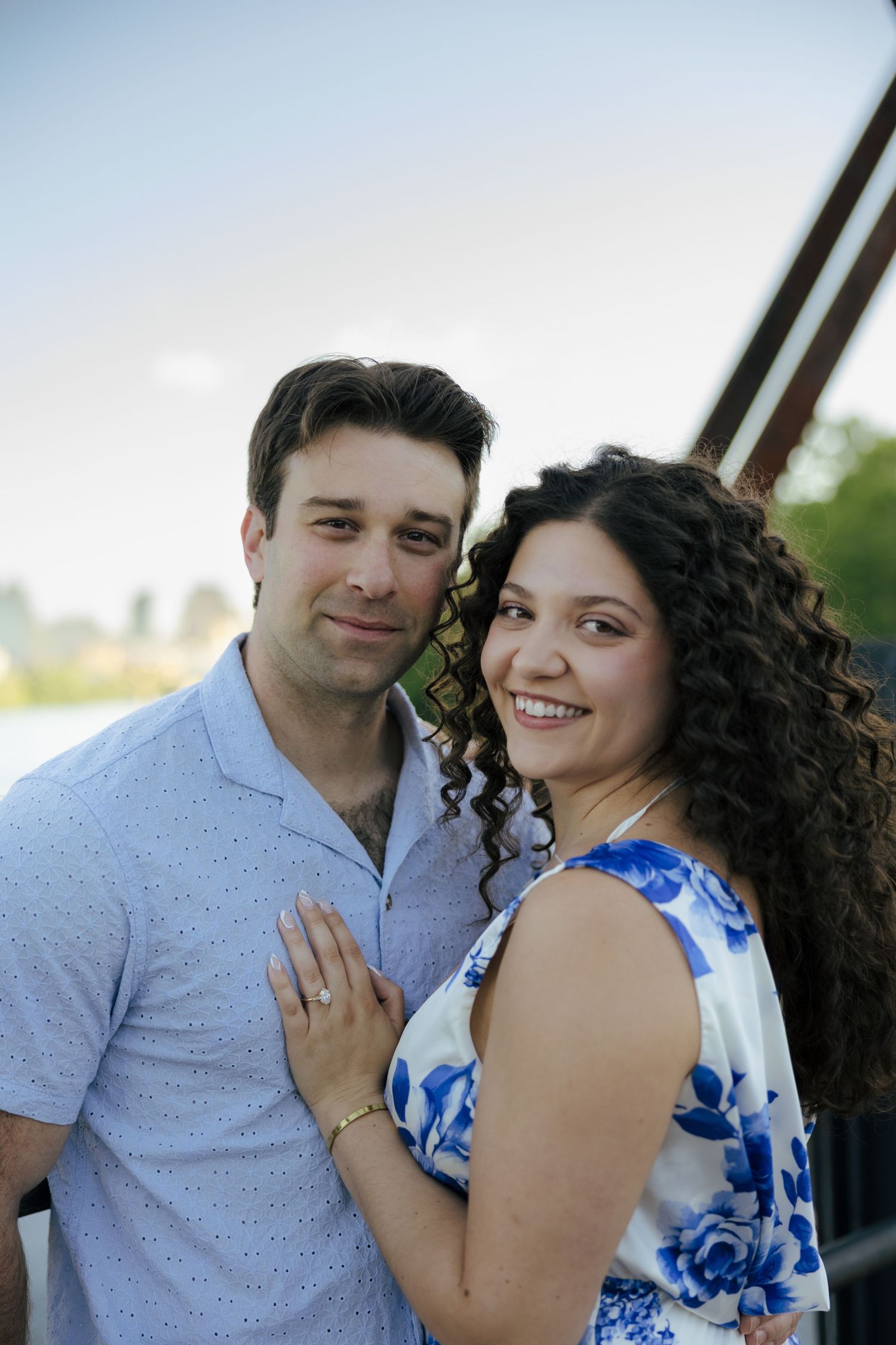 An engaged couple smiles together near a bridge at golden hour, the woman displaying her engagement ring.