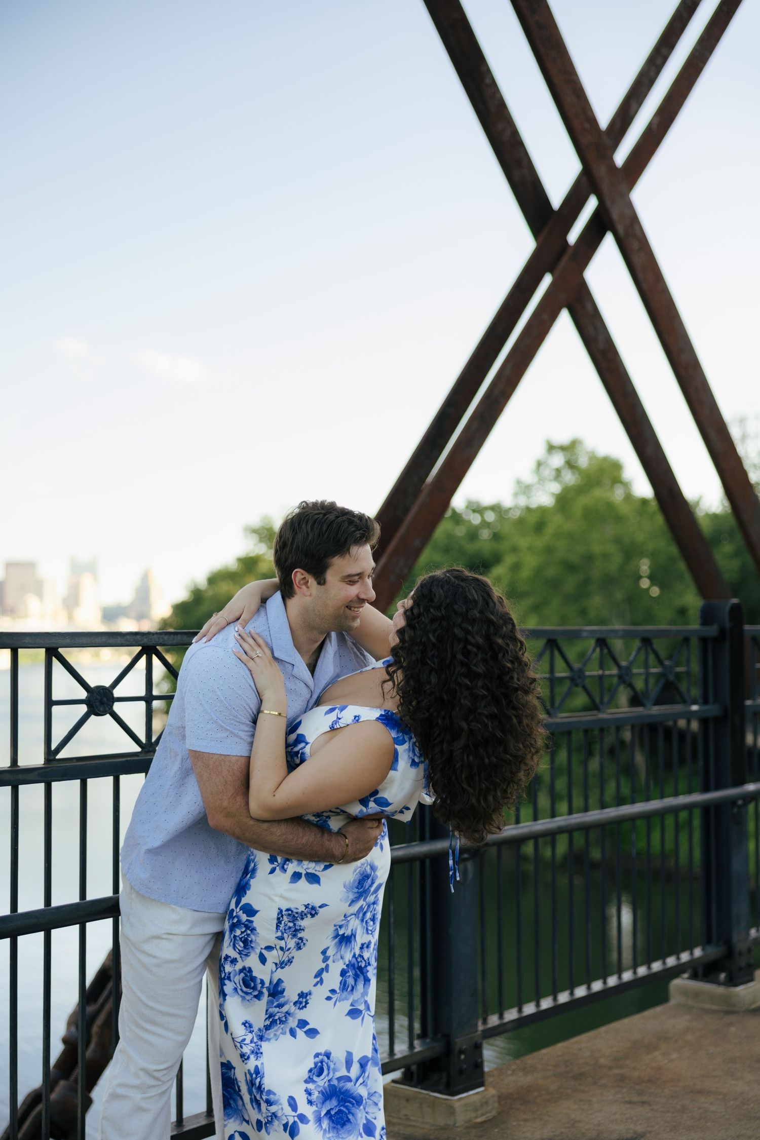 Couple sharing a romantic moment on a bridge with city skyline views, woman in blue floral dress.