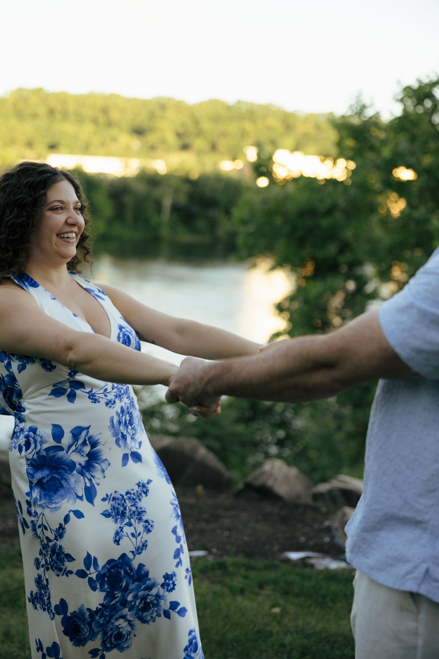 Joyful woman in blue floral dress holding hands with man outdoors near a scenic river at sunset.