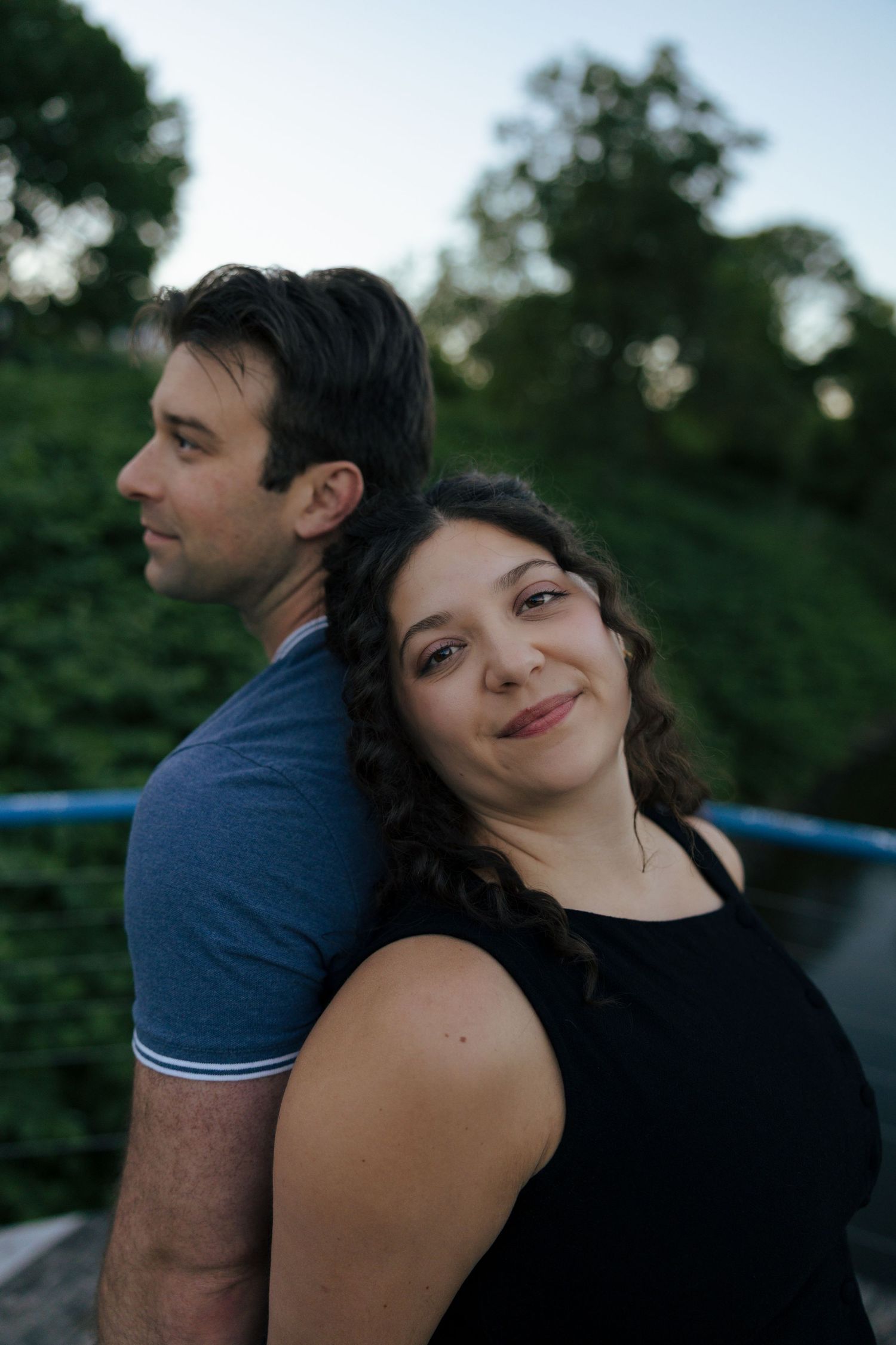 A couple poses back-to-back outdoors at dusk, the woman smiling warmly at the camera with green trees behind them.