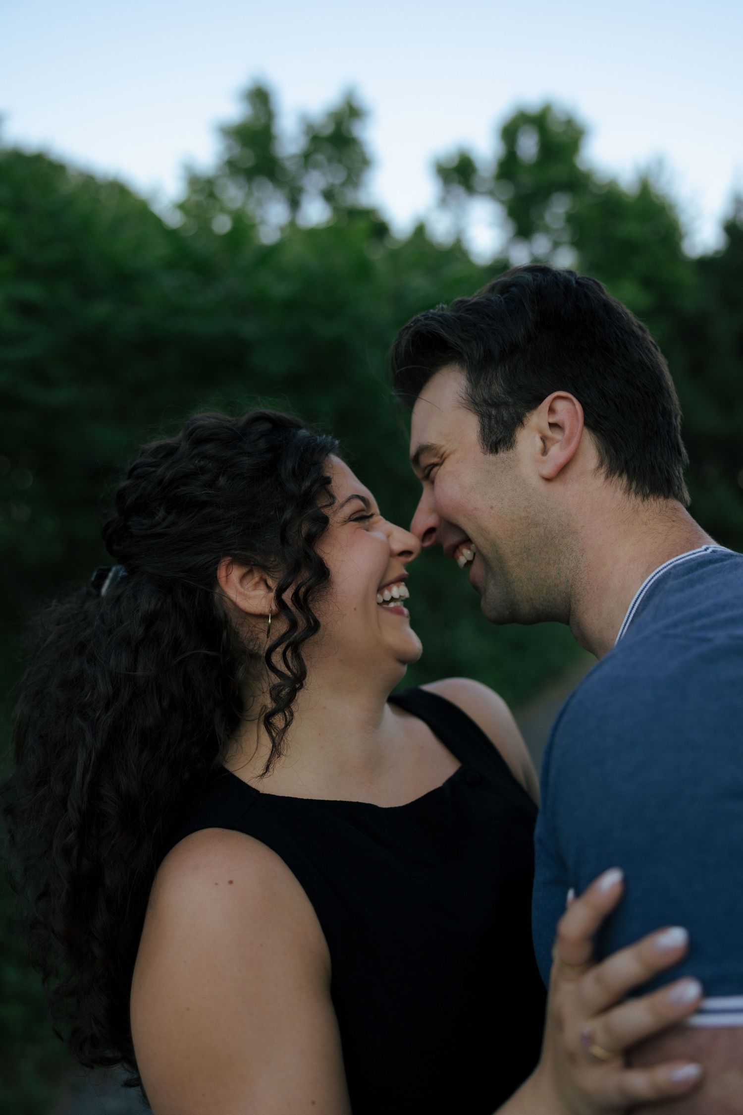 A smiling couple shares an intimate moment, nearly touching noses, surrounded by lush green trees at dusk.