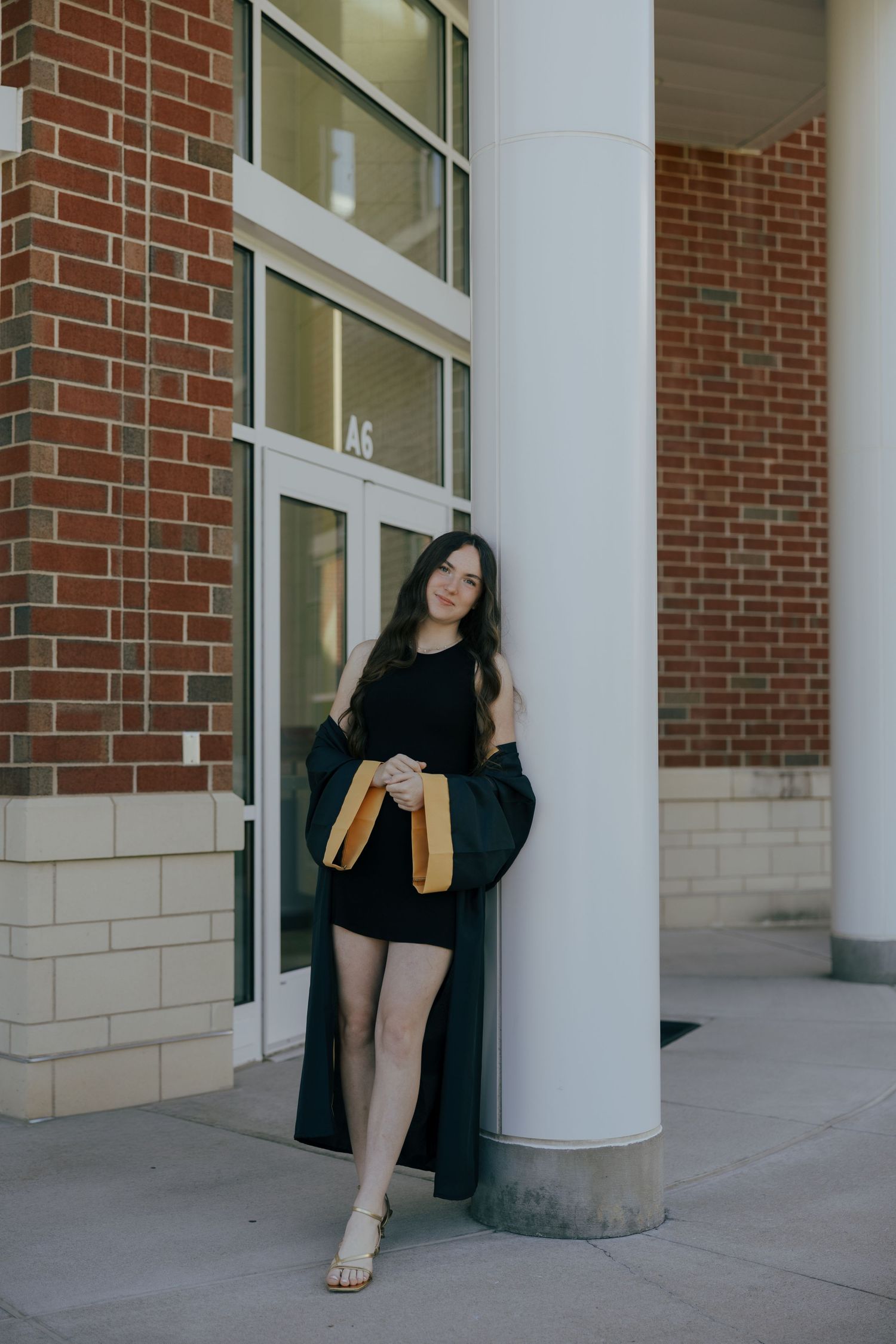 Young woman in black graduation gown holding gold tassel, posing by white column outside brick building.