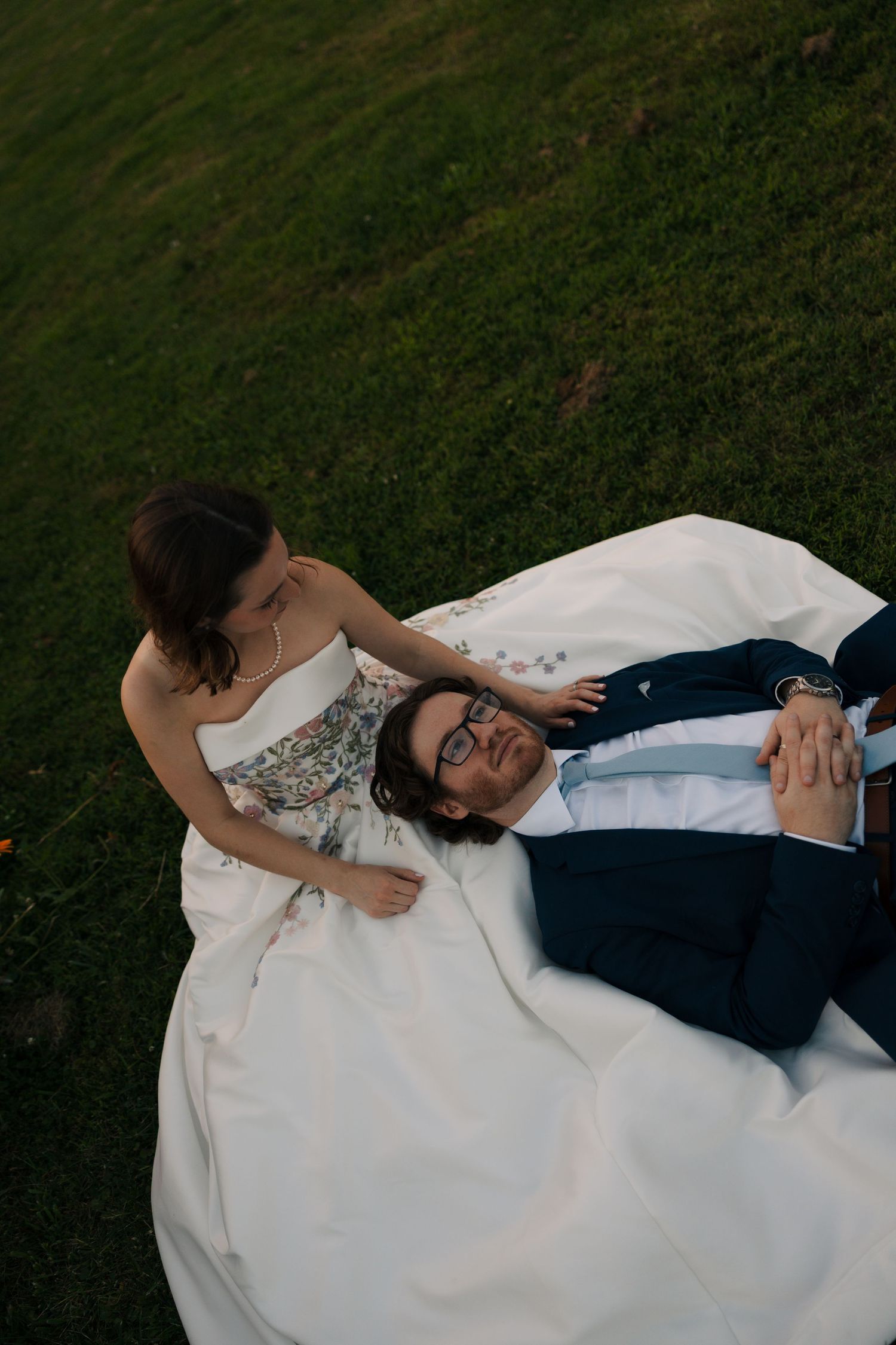 Groom in navy suit rests on bride's lap as she sits on grass in floral wedding gown at sunset.