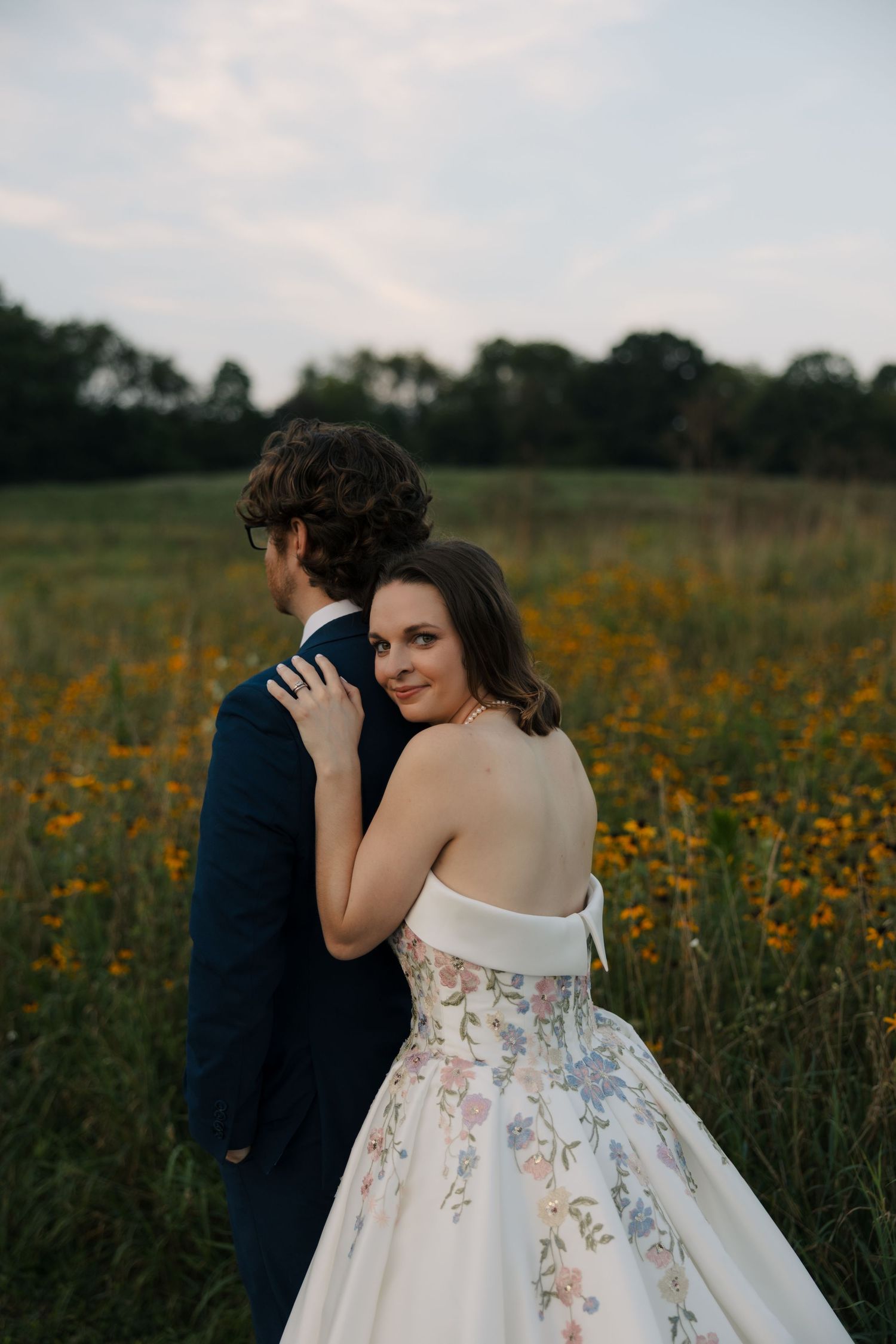 Bride in backless floral gown looks over shoulder while embracing groom in yellow wildflower field at sunset.