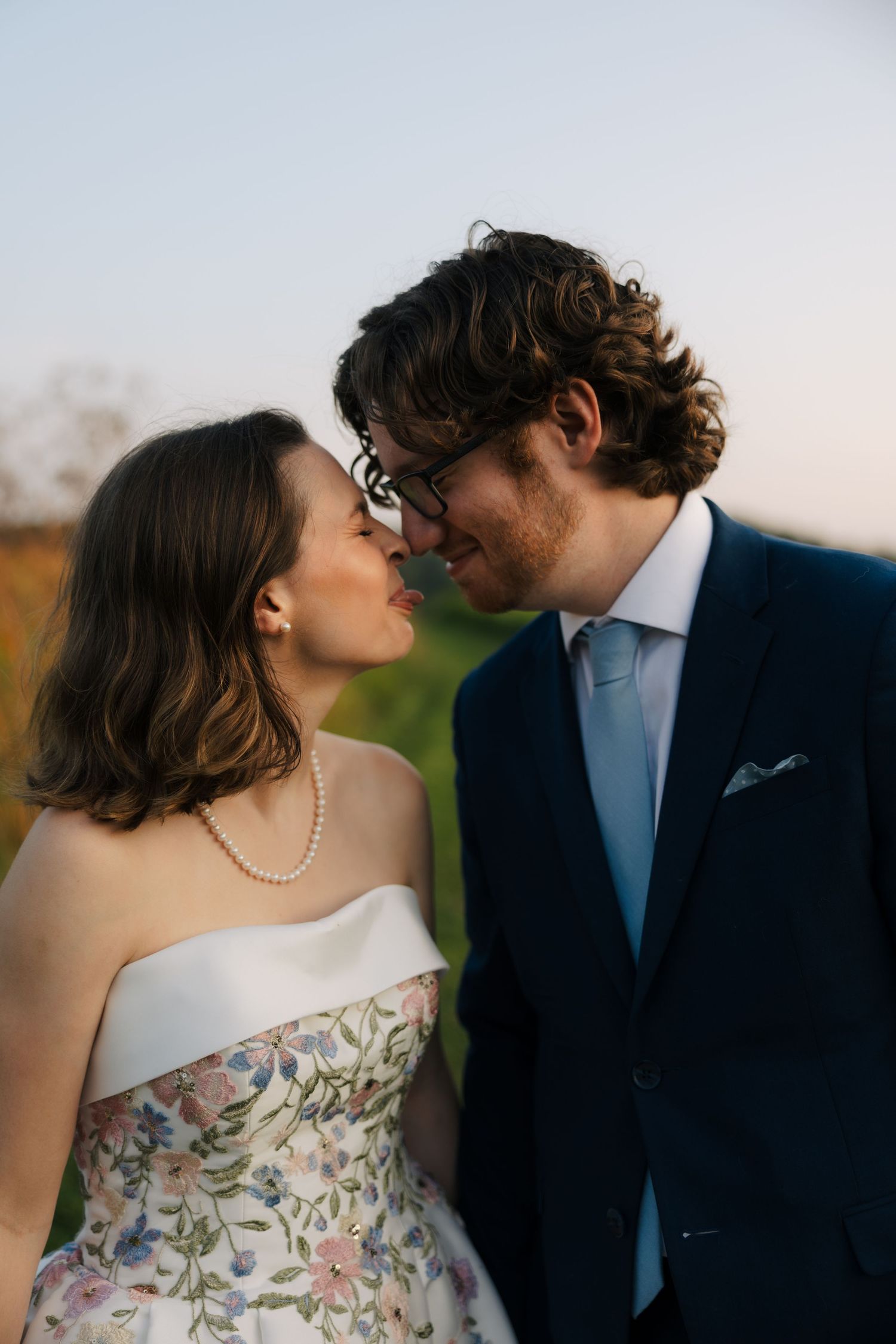 A bride and groom share a romantic moment, nearly kissing outdoors at golden hour during their wedding celebration.