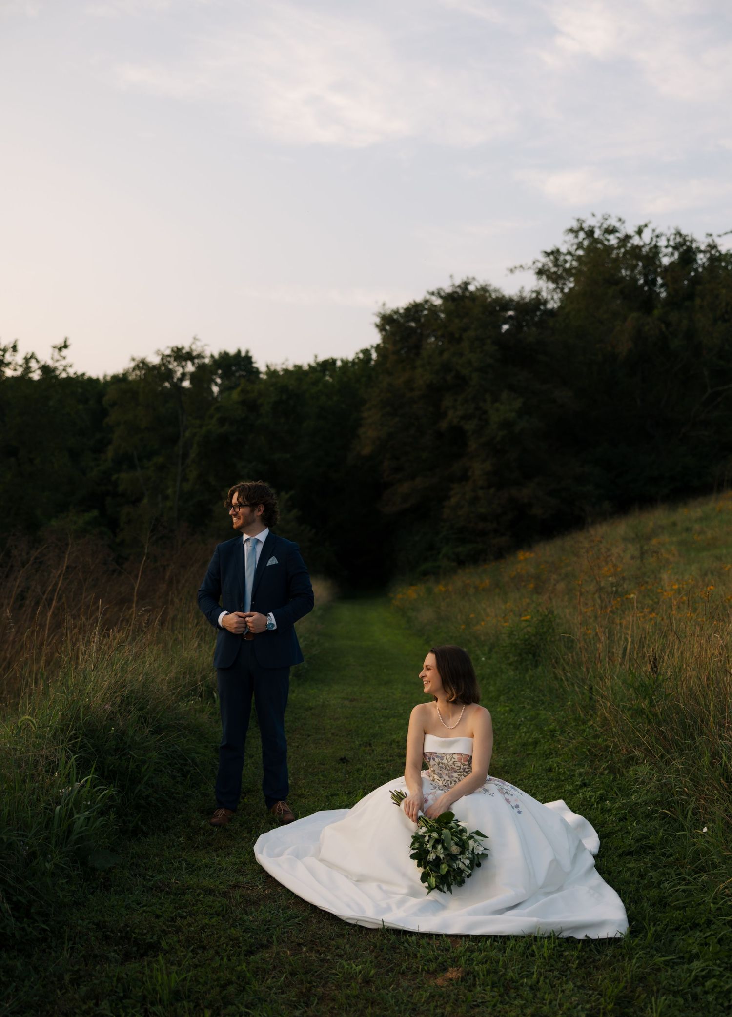 Bride in floral wedding gown sits on green hillside while groom in navy suit stands nearby at dusk.