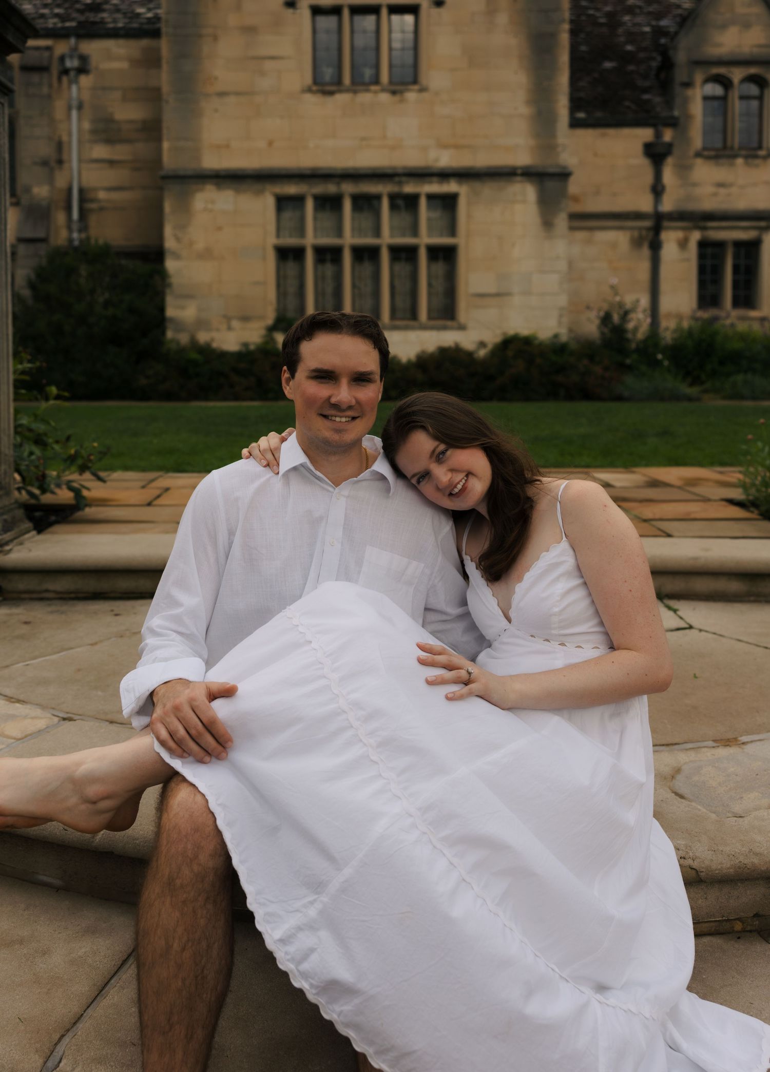 Smiling couple in white outfits sitting together on stone steps in front of a grand historic manor house.
