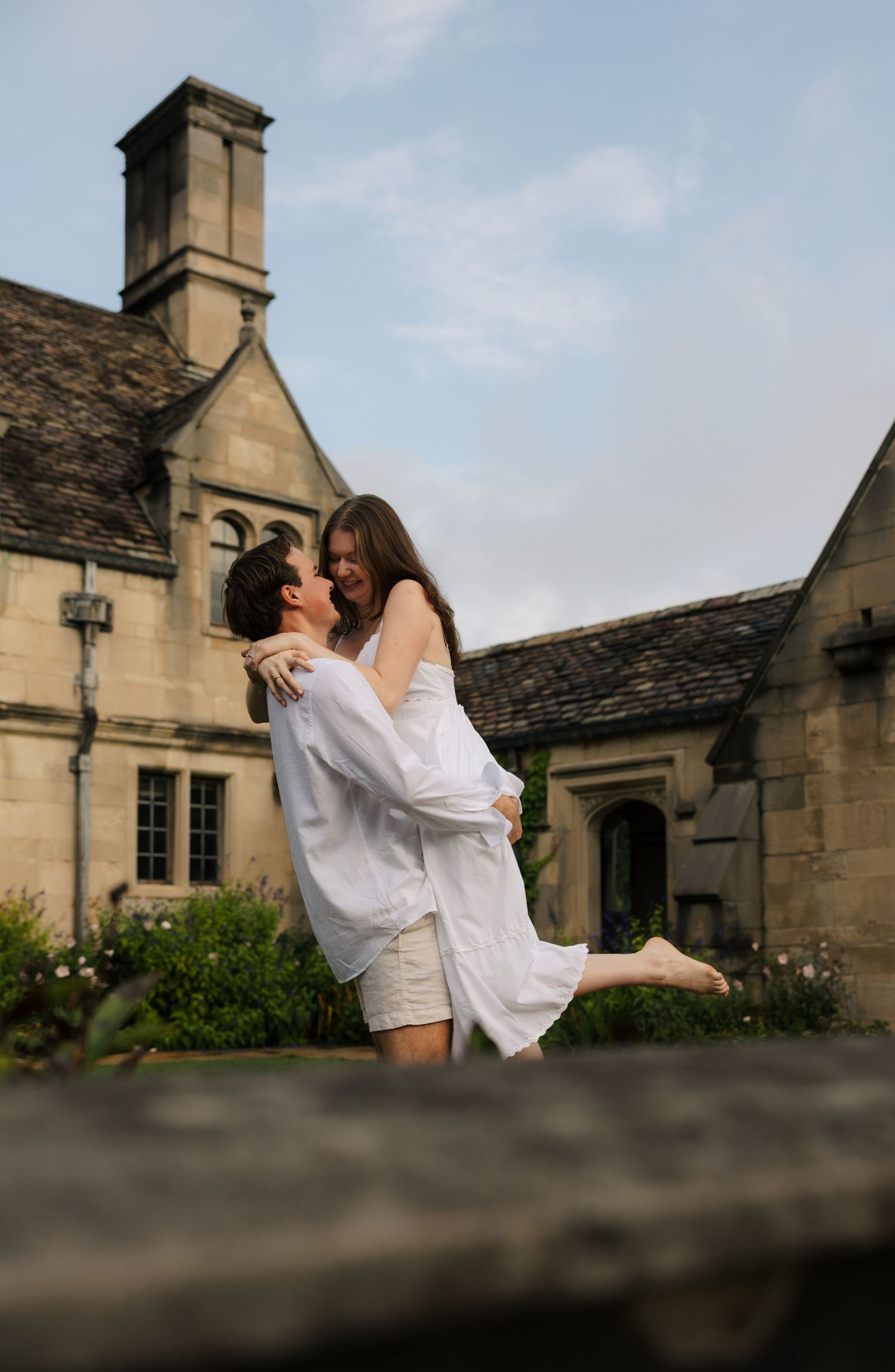 Joyful couple in white outfits embracing outdoors in front of a historic stone manor house.