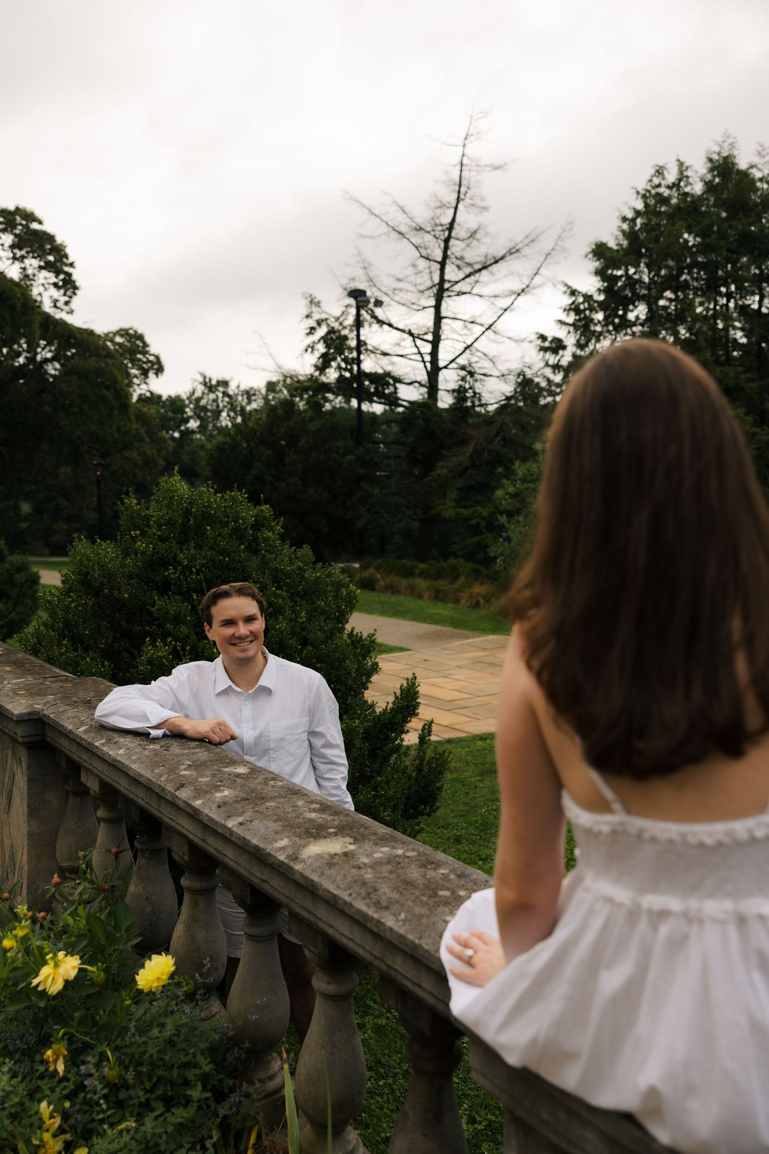 A smiling man in white shirt leans on stone balustrade, facing a woman in white dress in a lush garden setting.