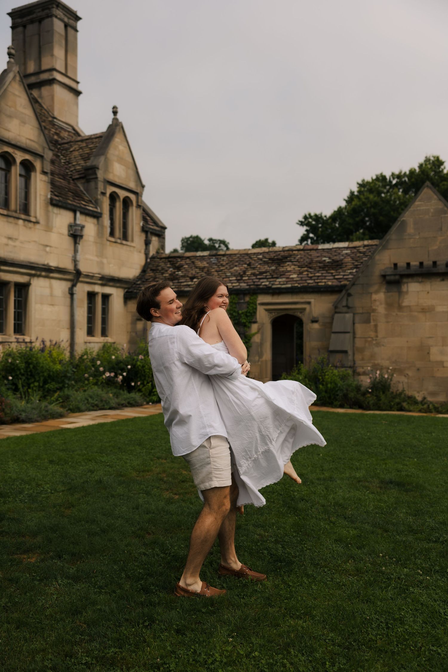A man joyfully lifts and spins a woman in a white dress on the green lawn of a beautiful stone estate.