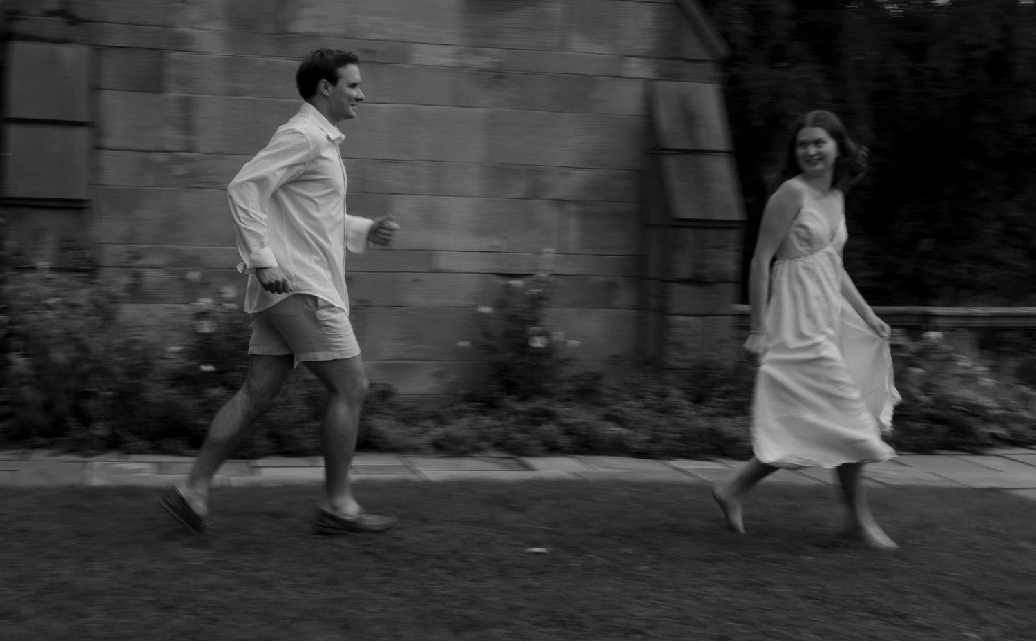 Black and white photo of a laughing couple running past a historic stone building.