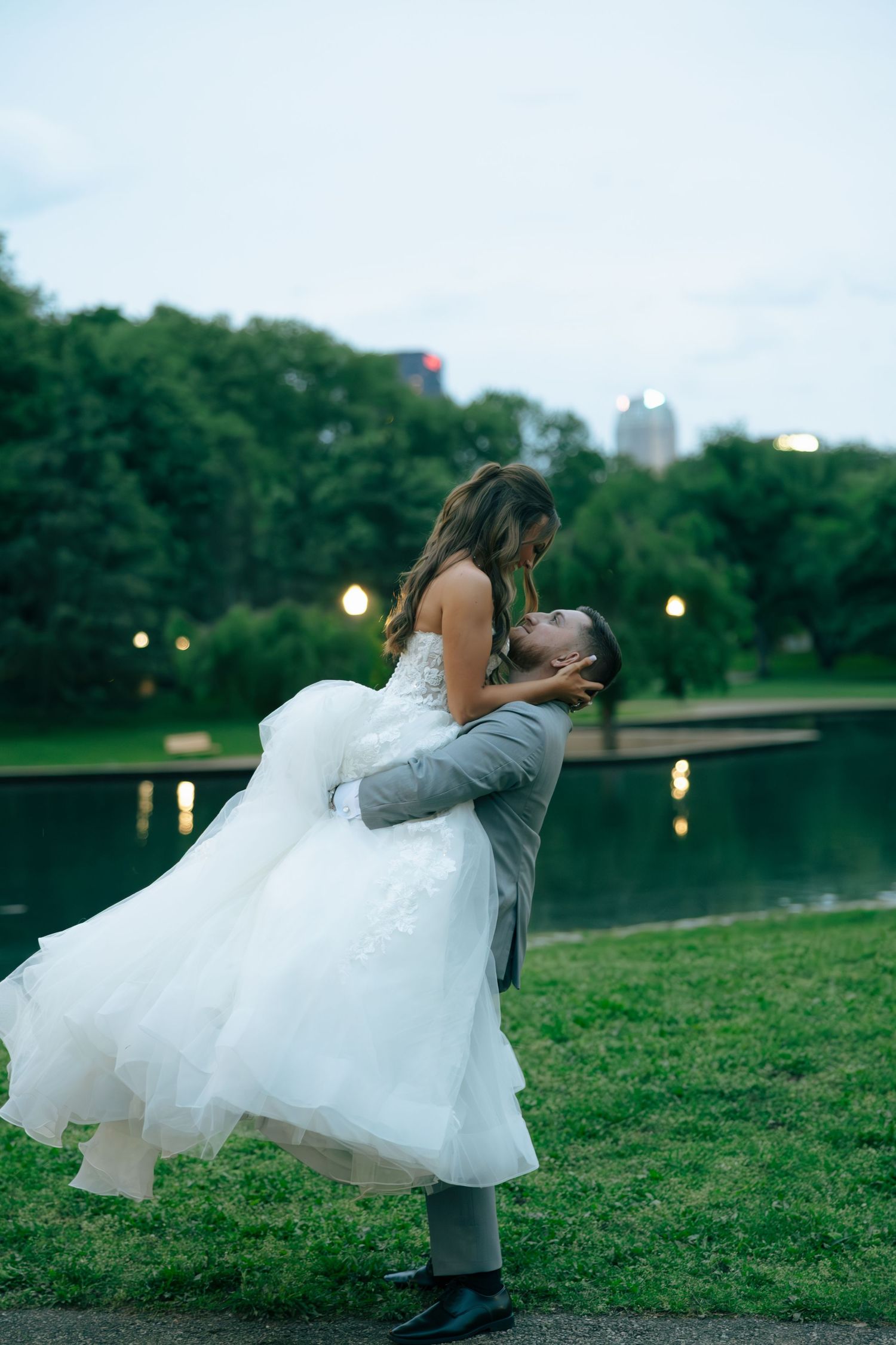 Groom lifts bride in white wedding dress by a pond in a lush green park at dusk.