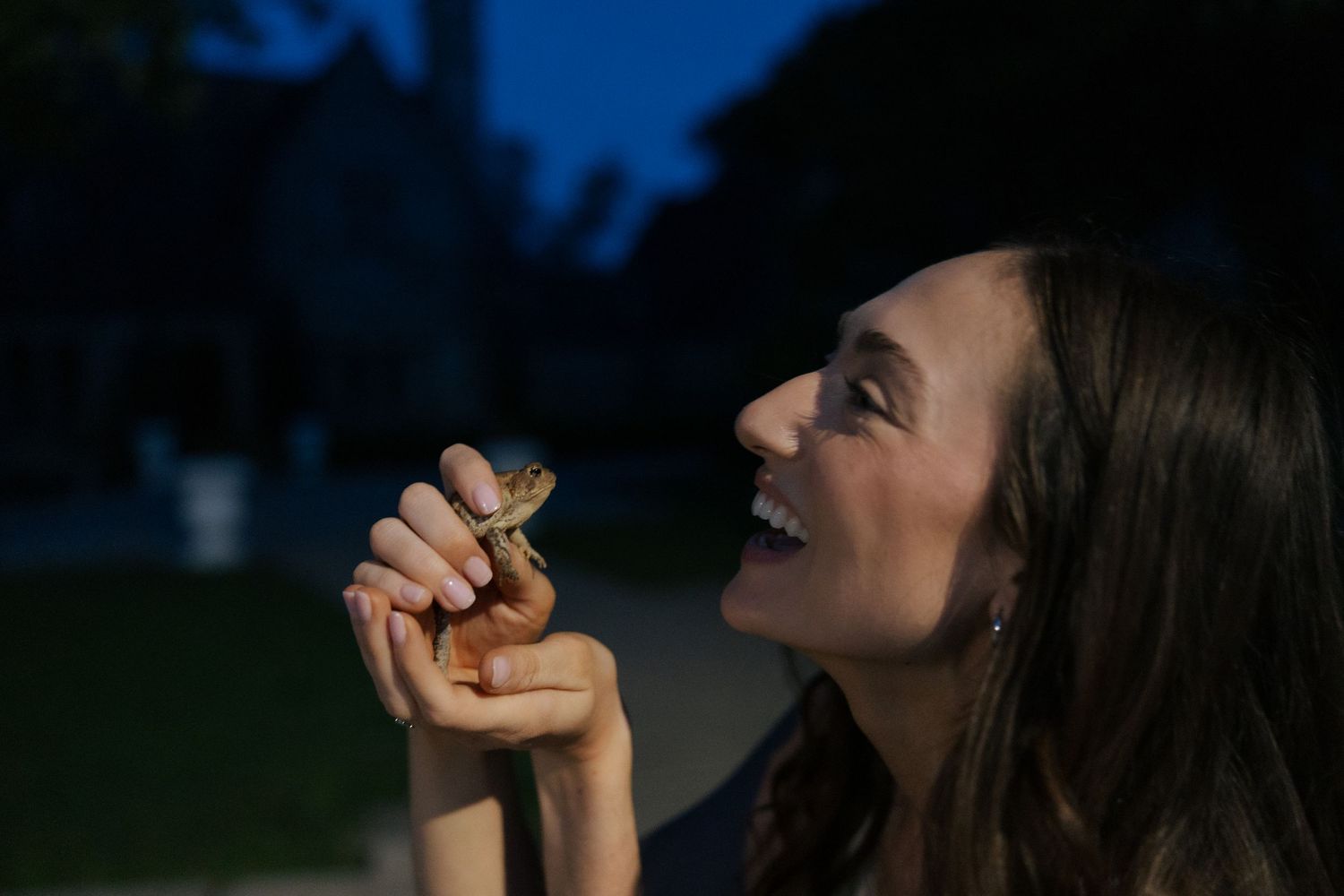 A delighted young woman smiles while holding a small lizard up close to her face at night.