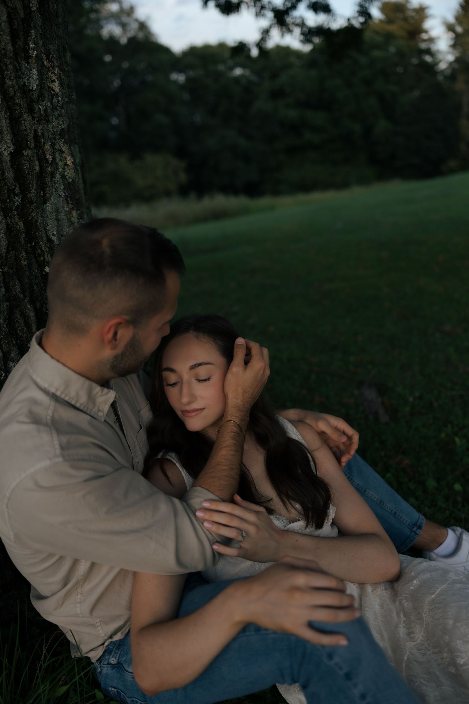 Couple sitting against a tree at dusk, man tenderly holding woman as she rests her head on his shoulder.