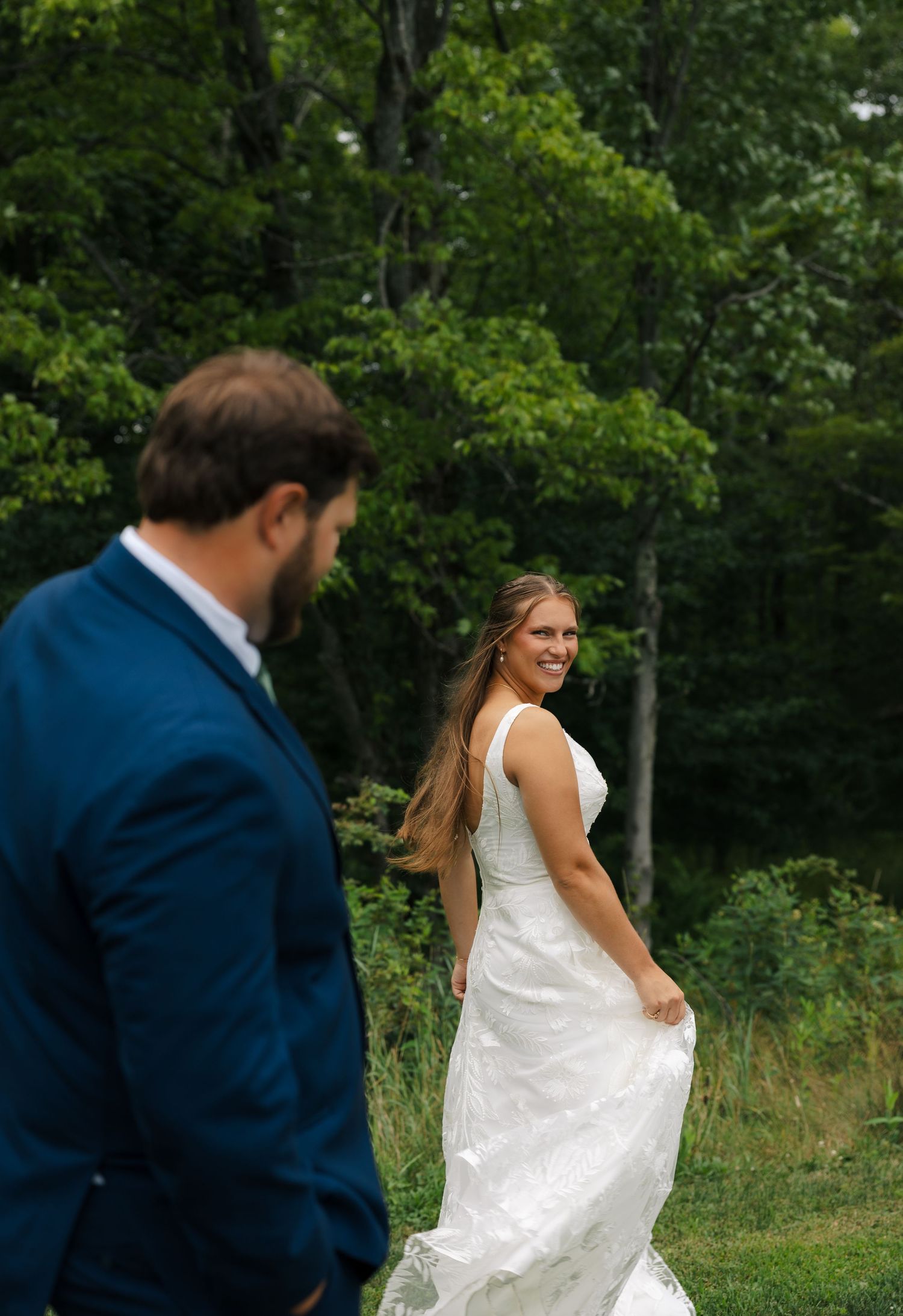 Bride in white gown smiles back at groom in blue suit during outdoor wedding ceremony surrounded by lush green trees.