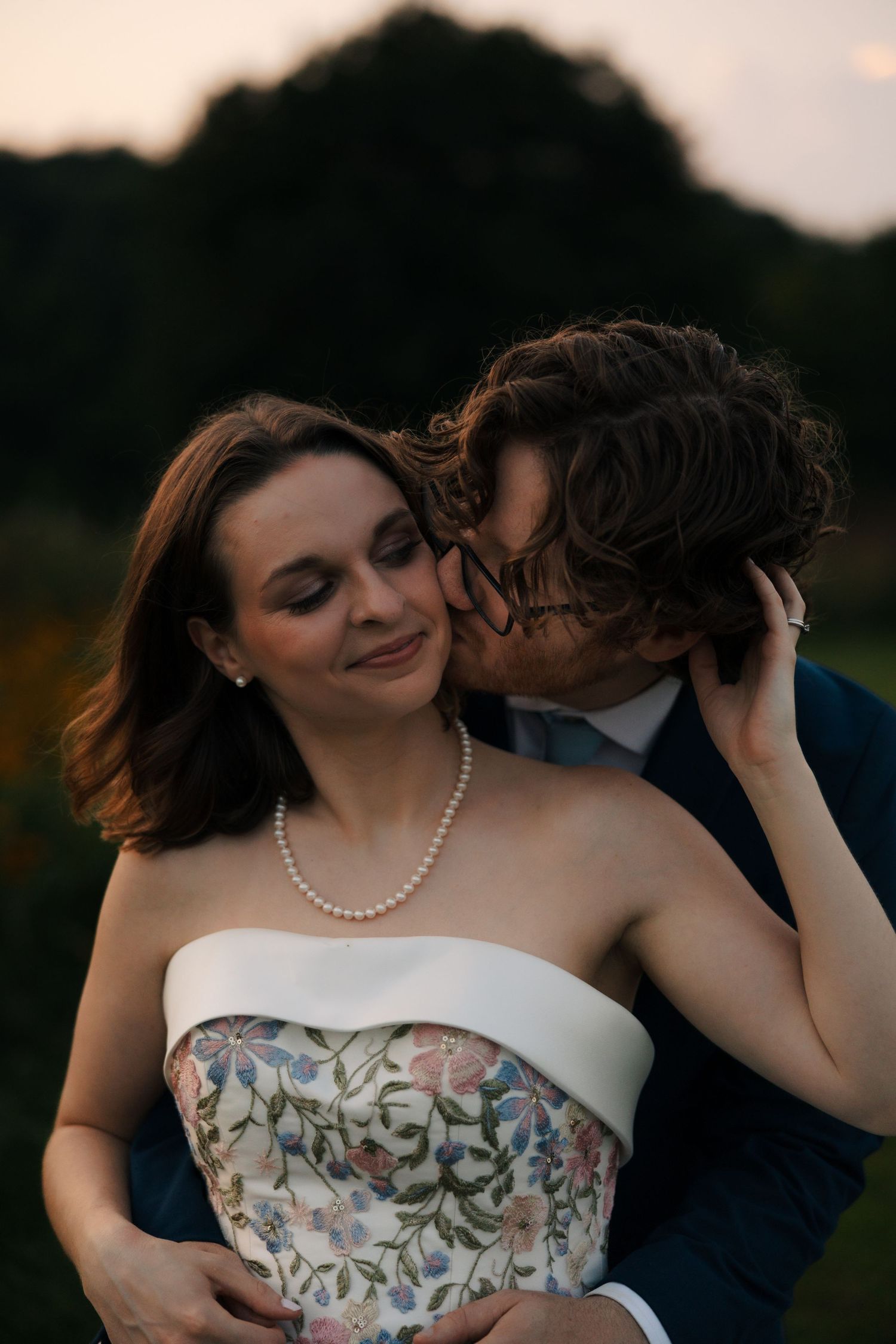 Groom in navy suit kisses bride wearing pearl necklace and strapless floral gown at twilight.