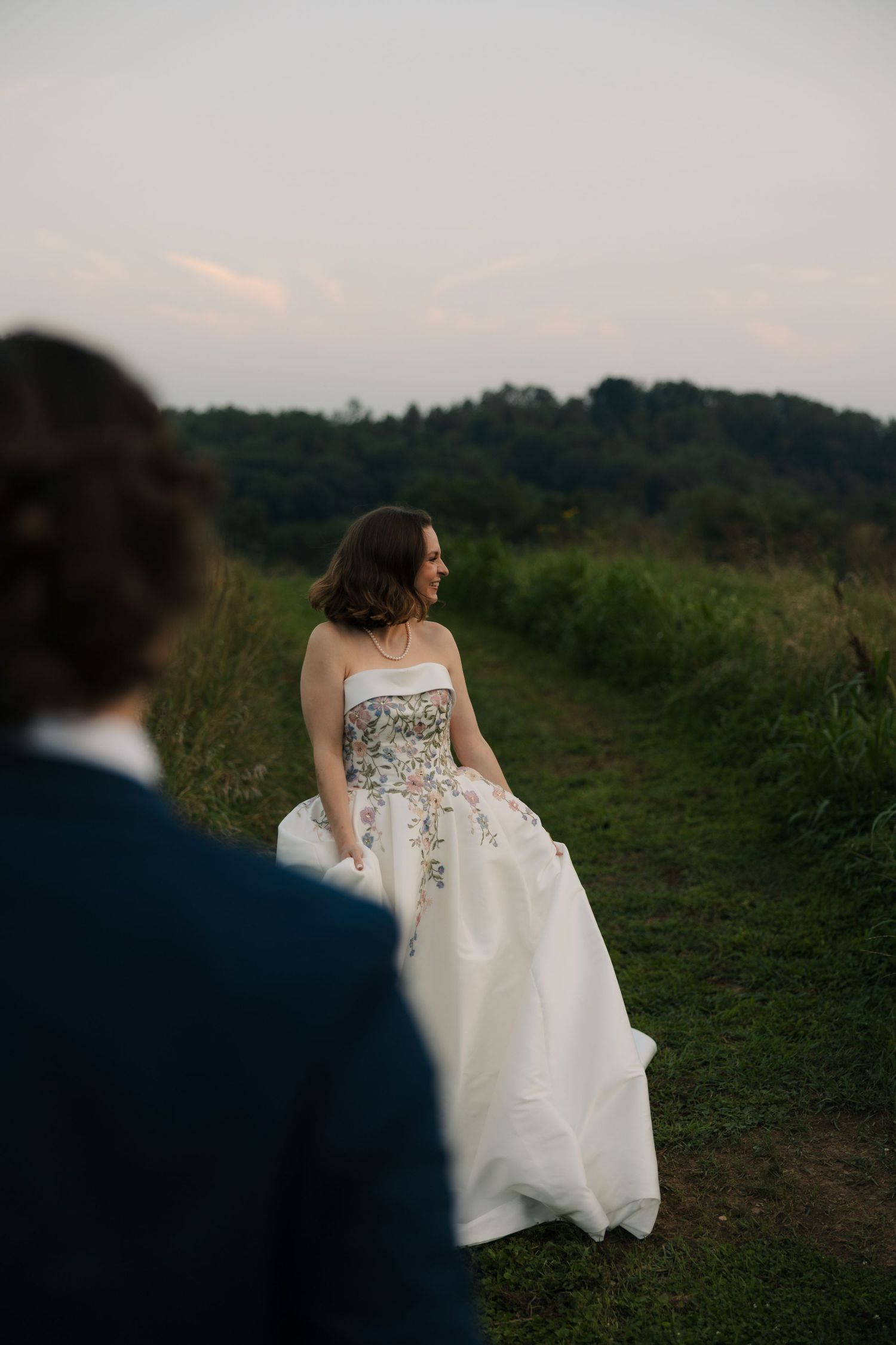 Bride in floral strapless wedding gown walks path toward groom in navy suit at golden hour.