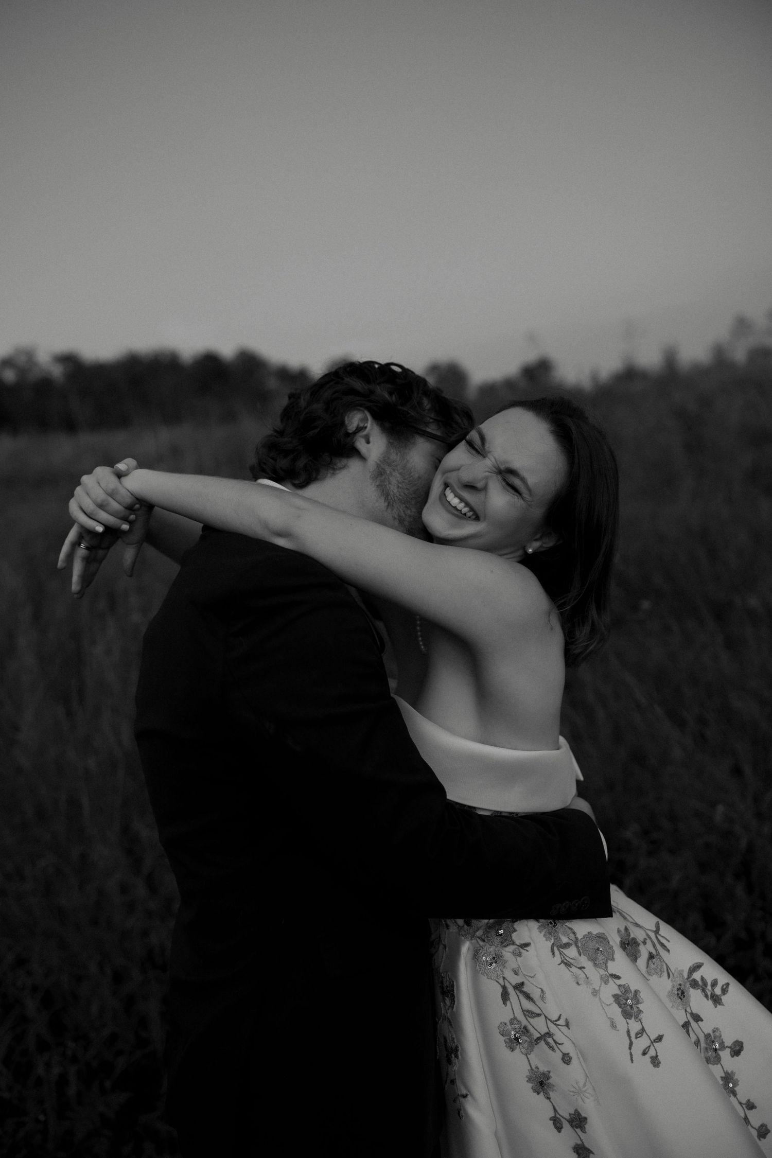 Black and white photo of smiling bride embracing groom in field during romantic outdoor wedding portrait.