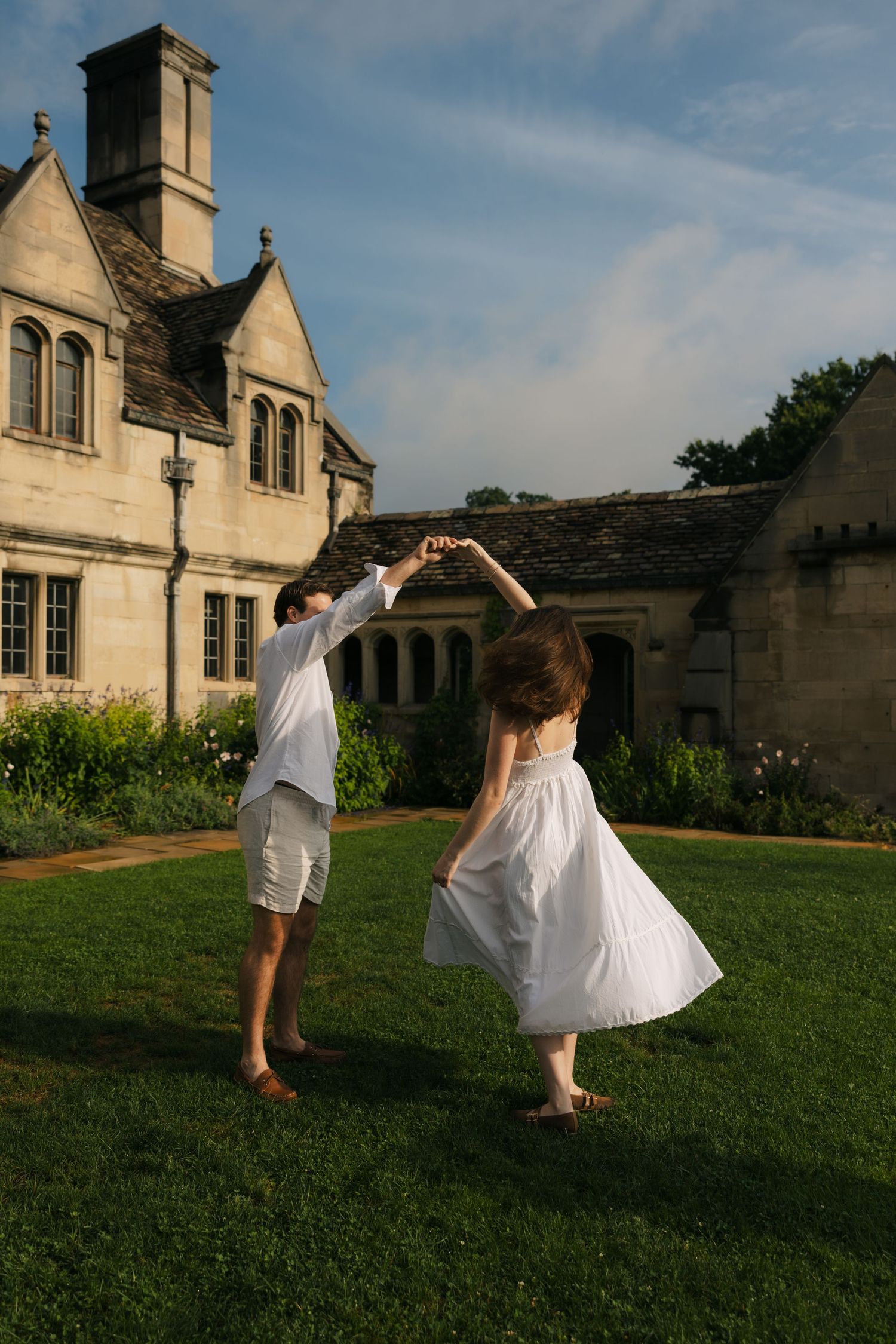 Couple dancing barefoot on green lawn before a historic stone manor house on a sunny day.