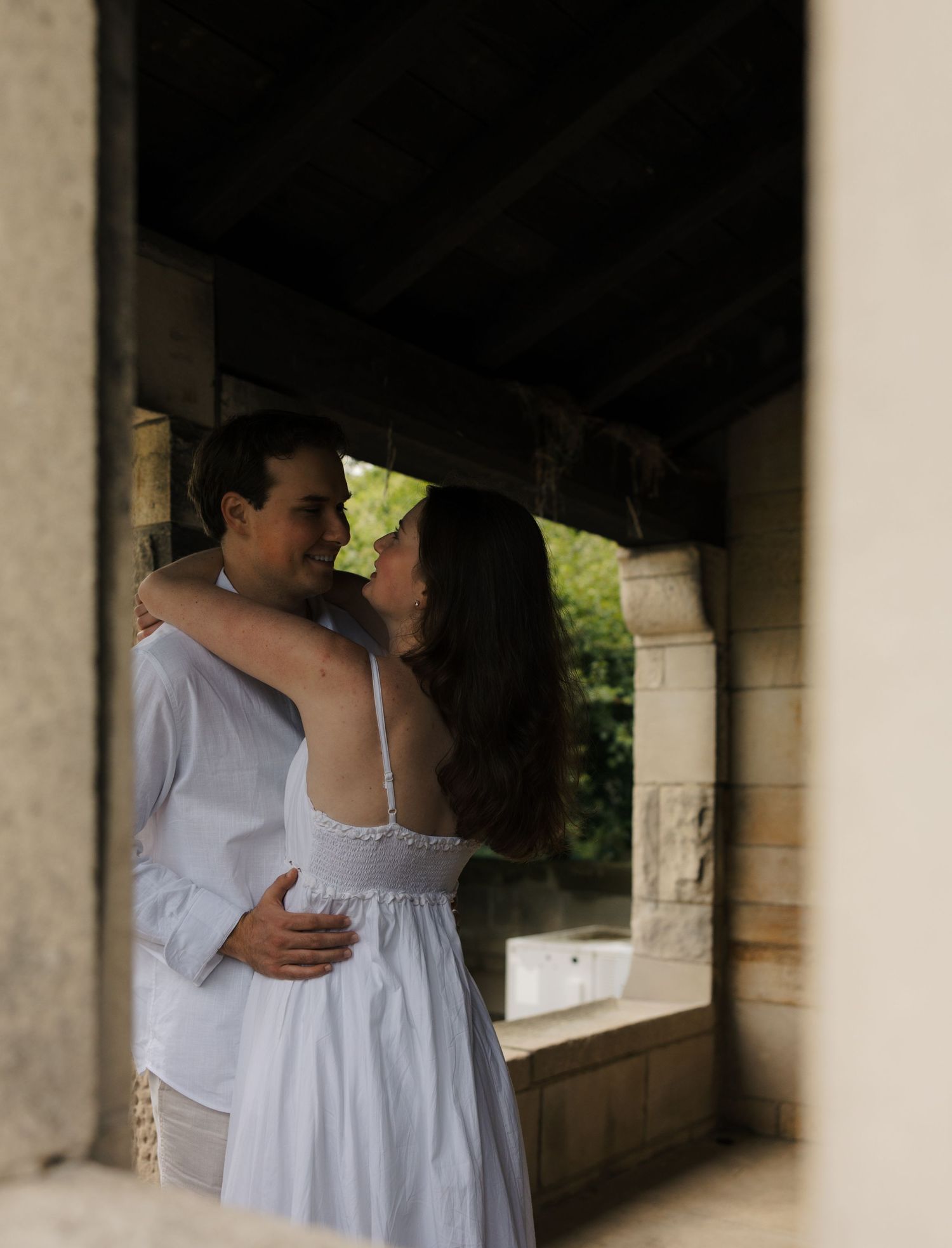 Romantic couple in white embracing under stone archway in soft light.