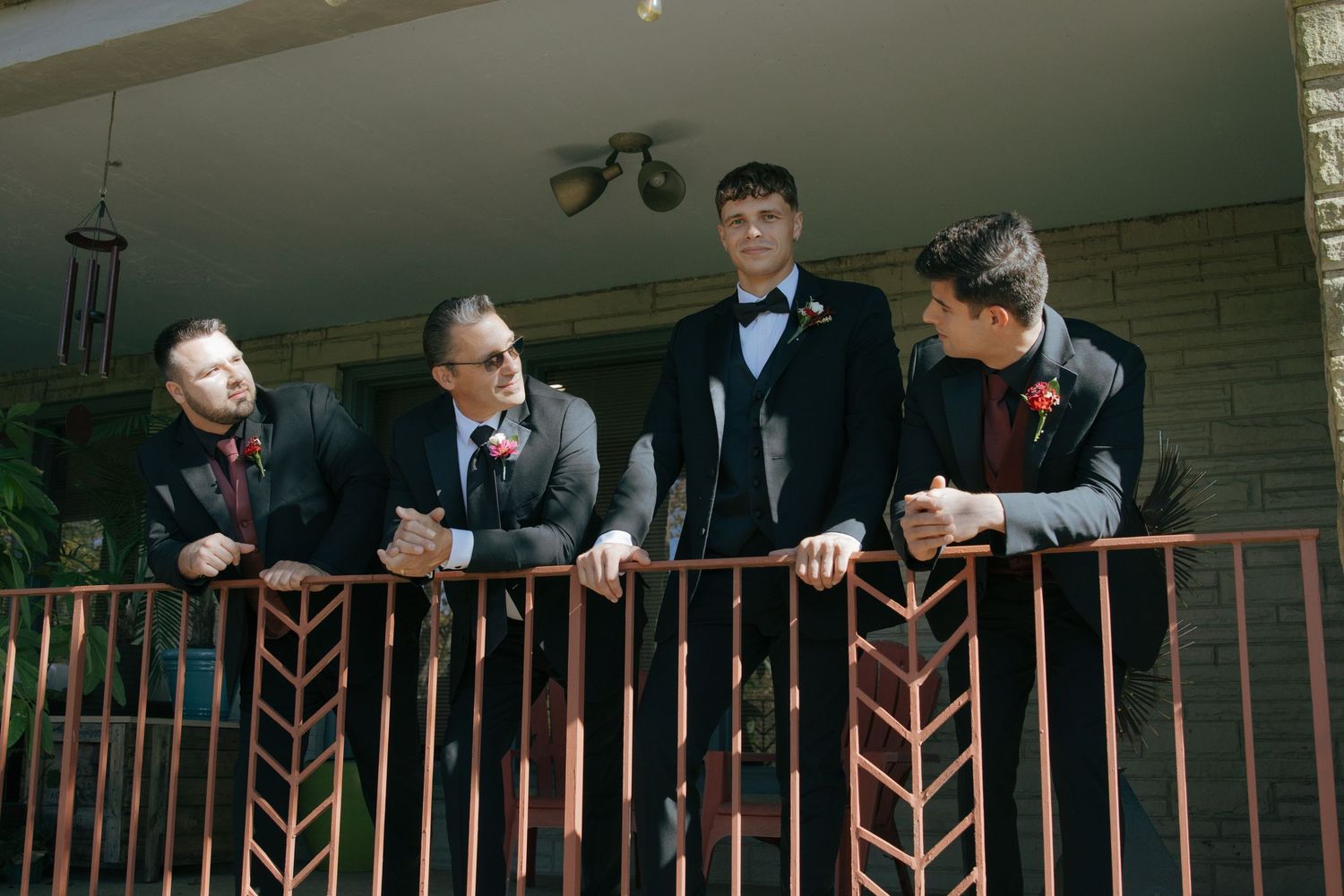 Four men in formal suits and boutonnieres stand on a decorated balcony, likely at a wedding celebration.