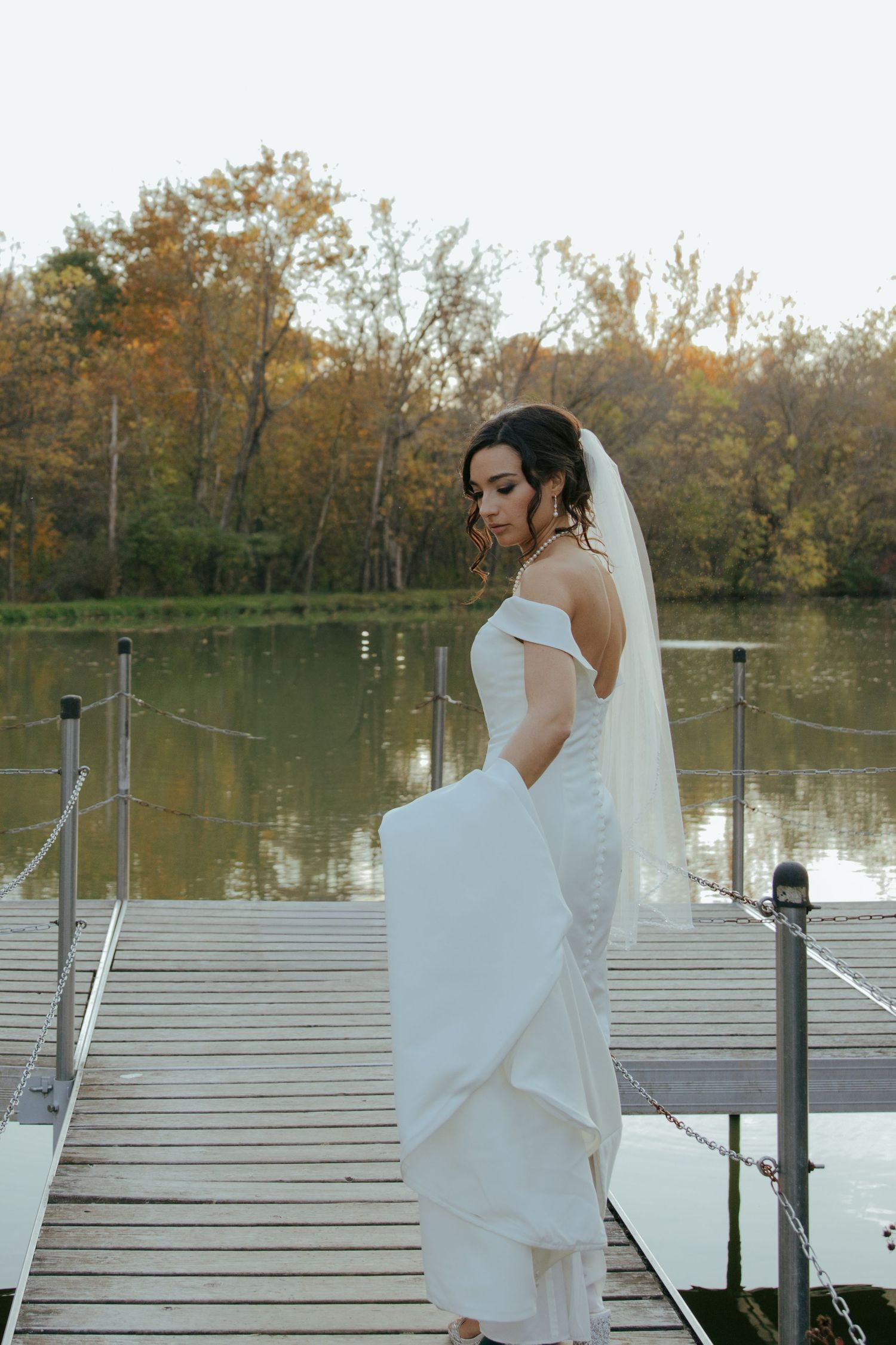 Elegant bride in white gown and veil standing on a wooden dock by a serene autumn lake.
