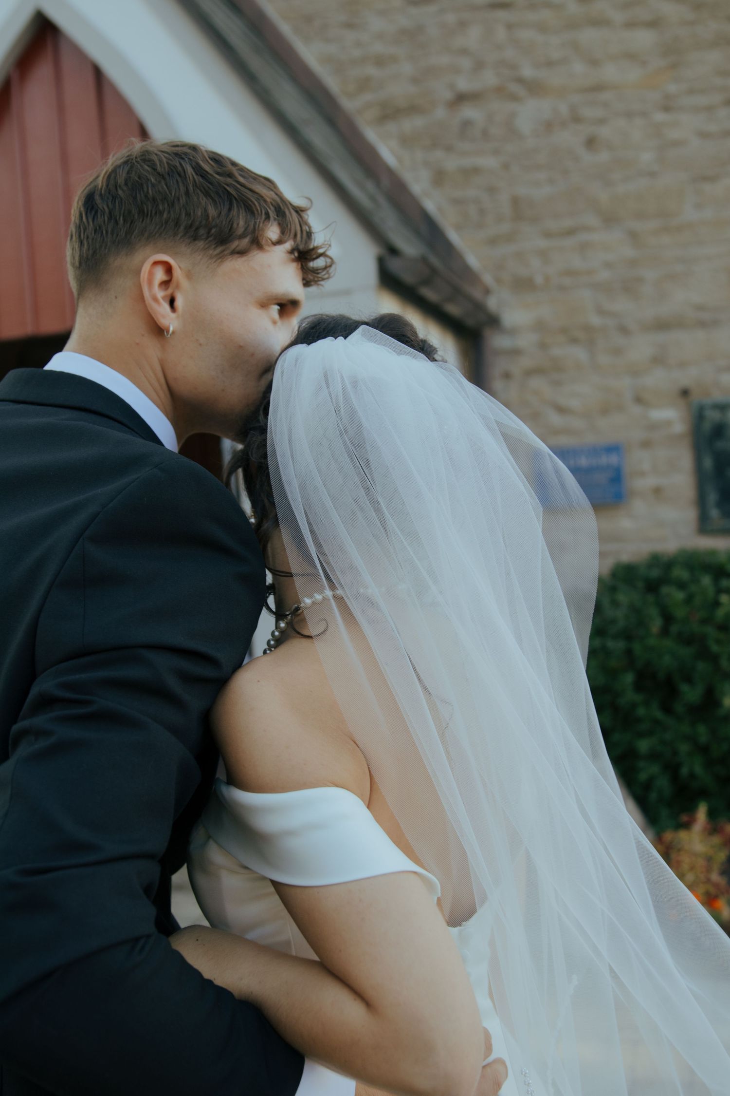 Groom in black suit kisses bride in off-shoulder white gown and veil outside a rustic stone church.