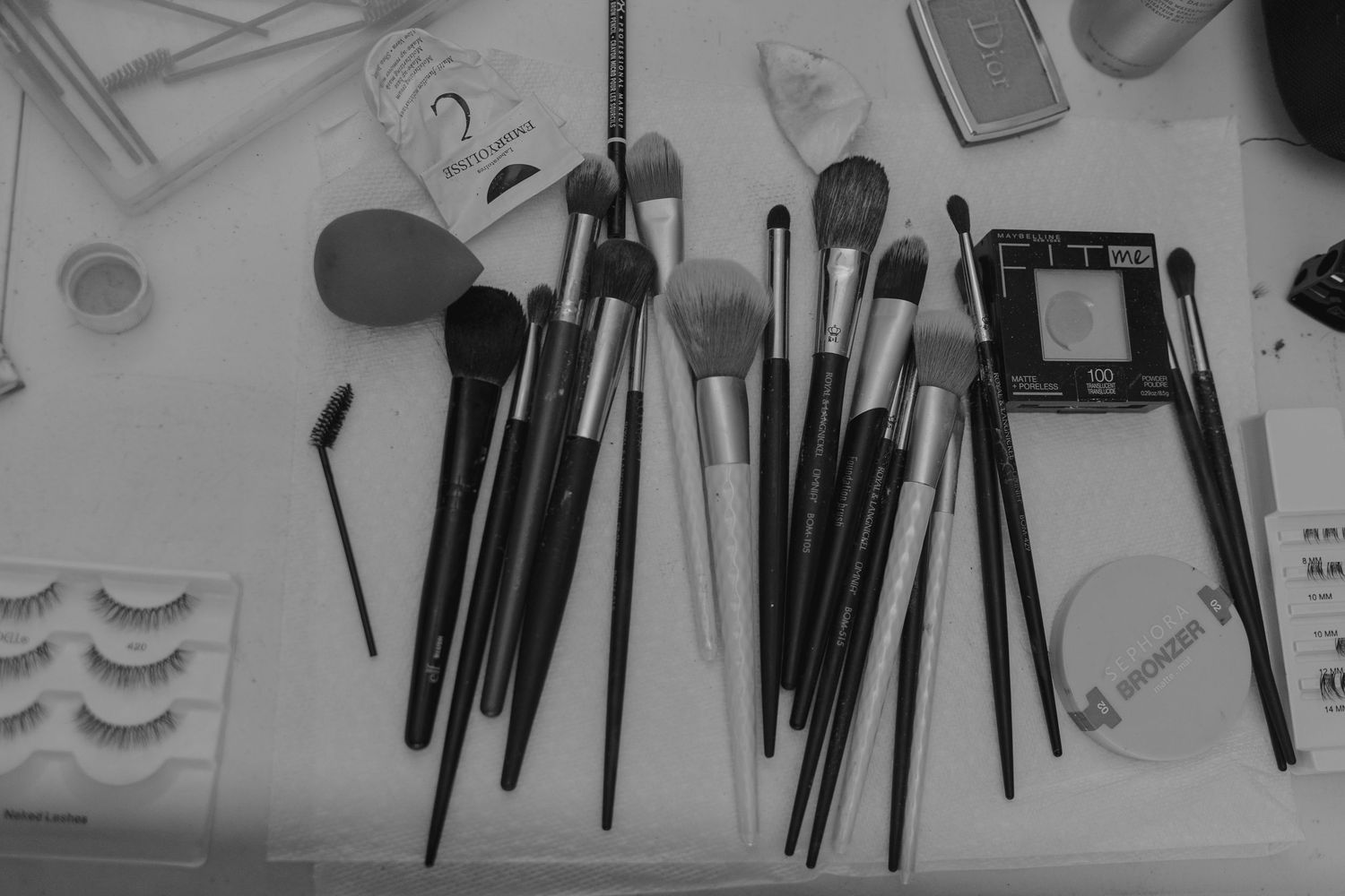 Black and white photo of assorted makeup brushes and beauty tools arranged on a table with false lashes and cosmetics.