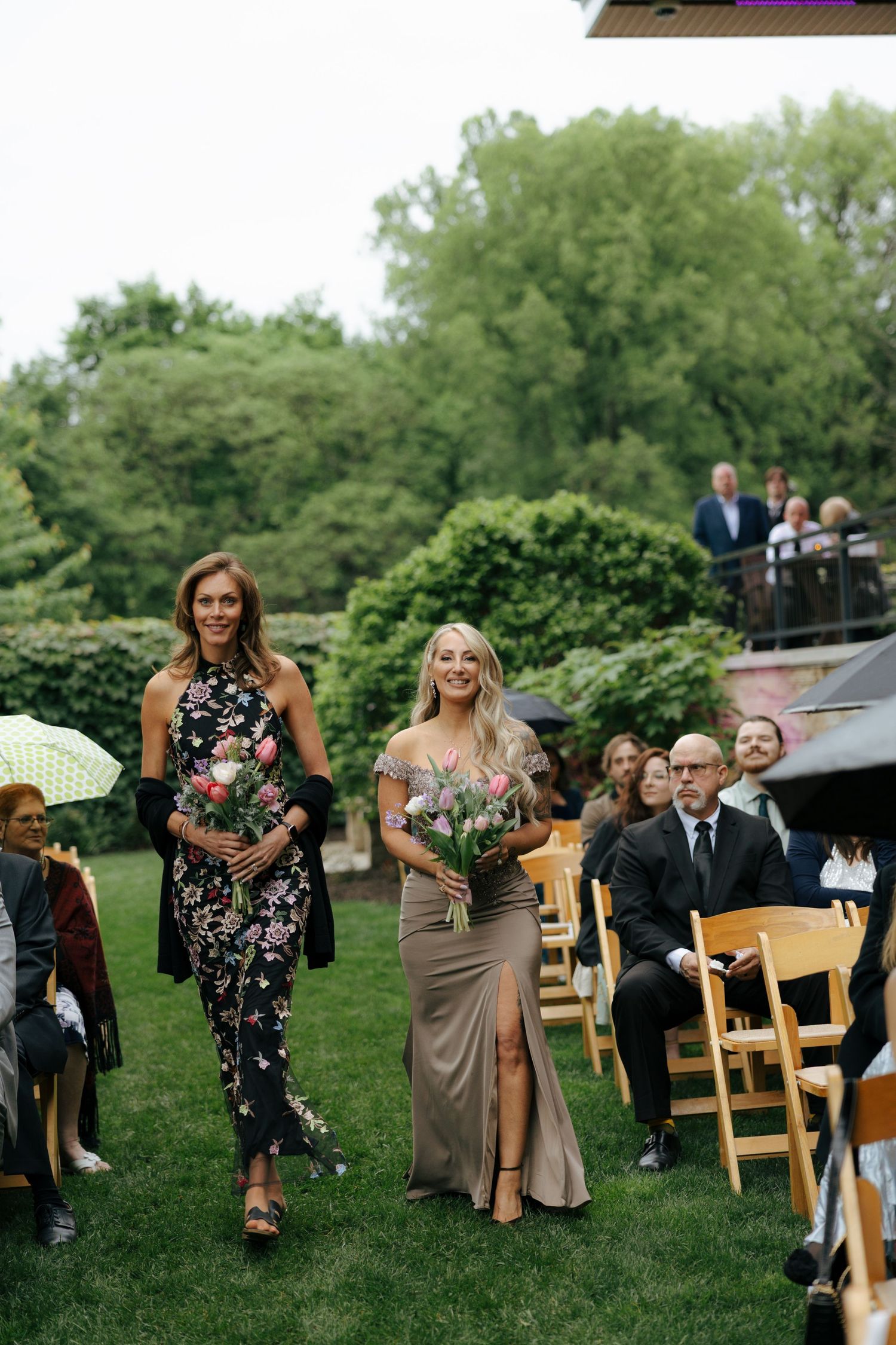 Two bridesmaids walk down the aisle at an outdoor garden wedding ceremony, one in floral dress, one in gold gown.