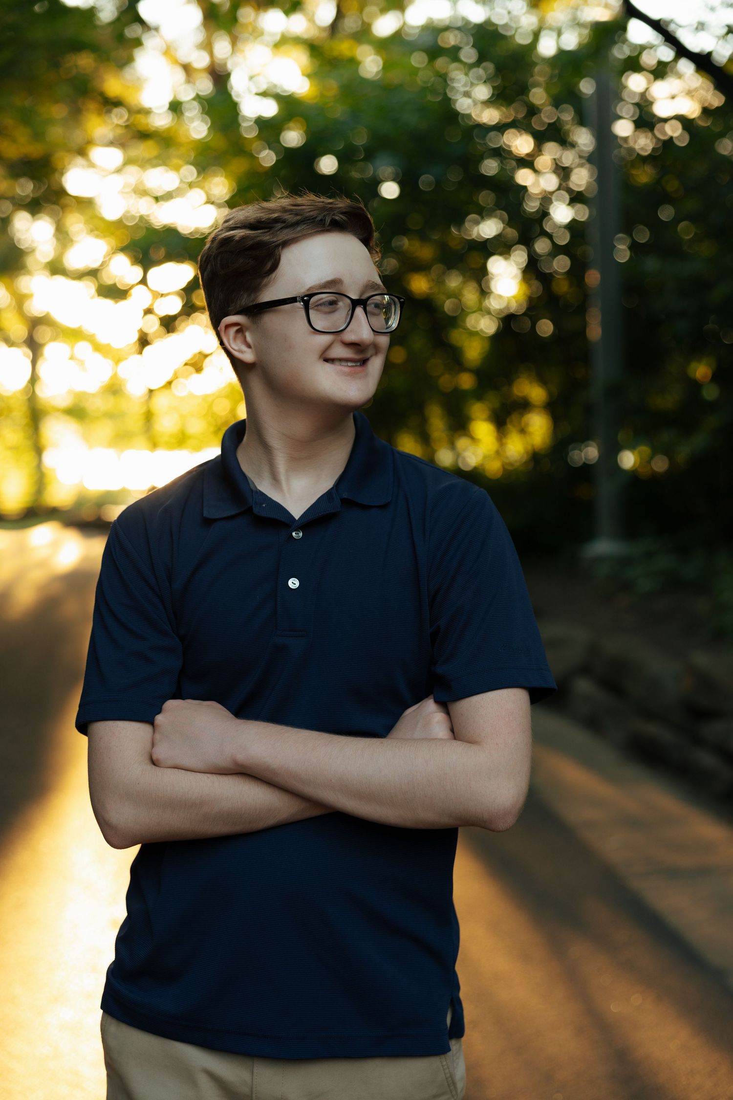 Teen boy in navy polo with arms crossed smiling outdoors during golden hour sunset in a park.