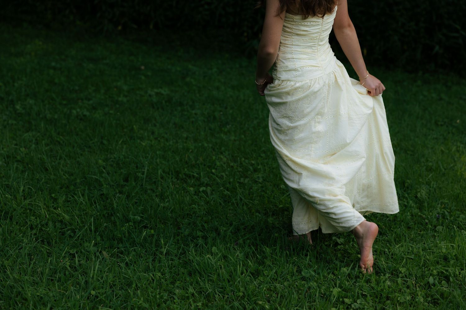Barefoot woman in flowing white wedding dress walking on lush green grass.