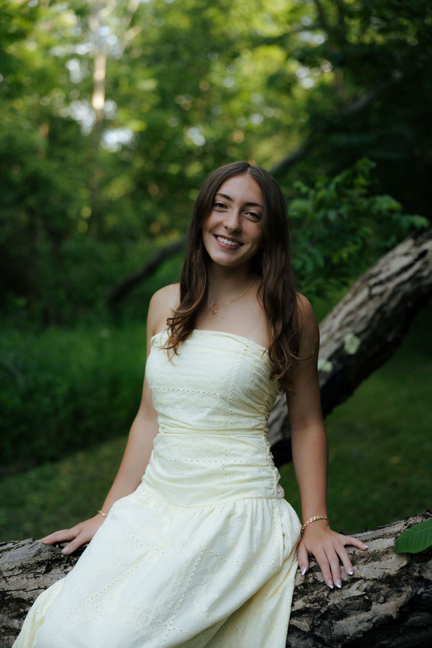 A smiling young woman in a pale yellow strapless dress sits on a fallen log in a forest.