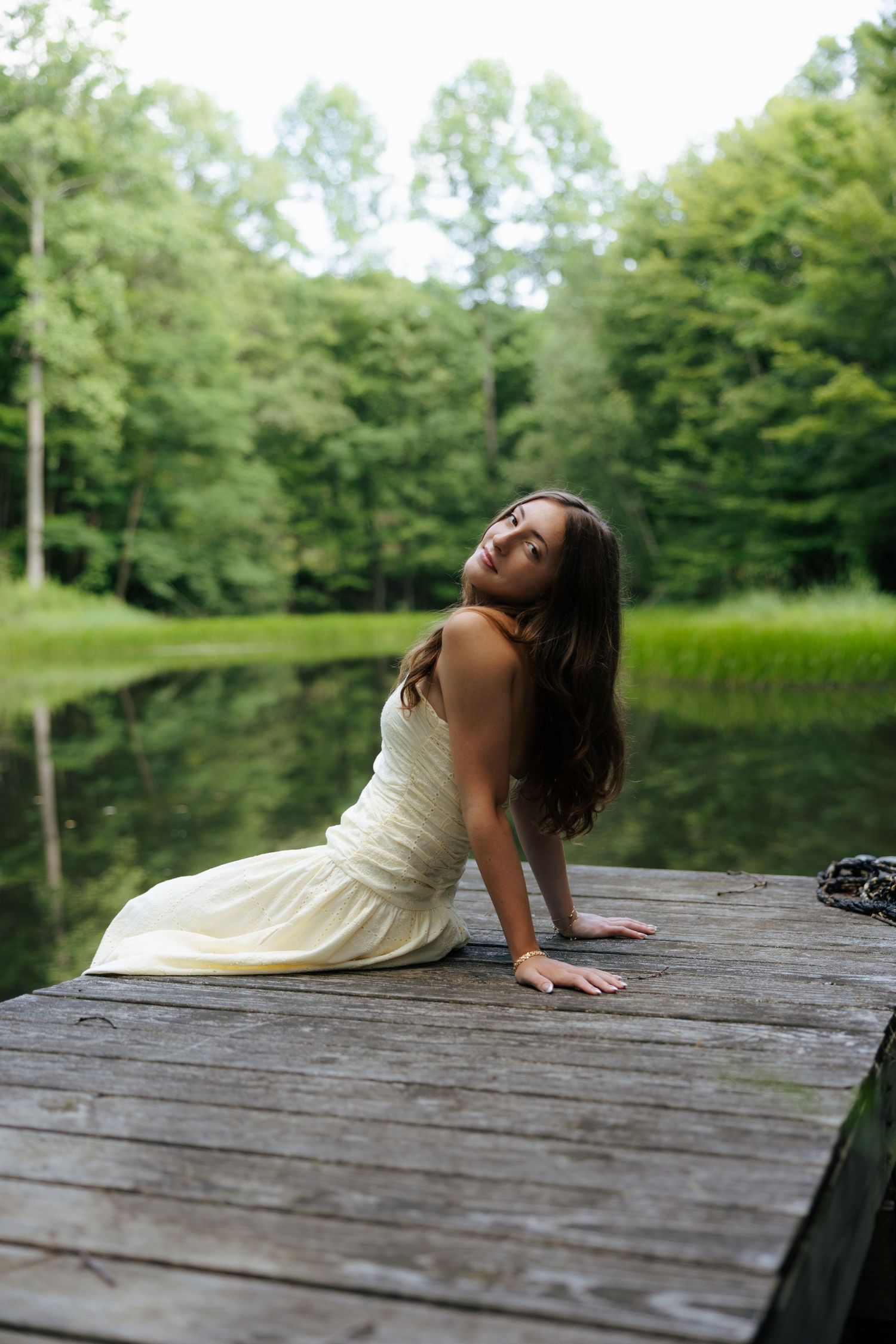 Young woman in white dress sitting on wooden dock tilting head back by tranquil forest lake.