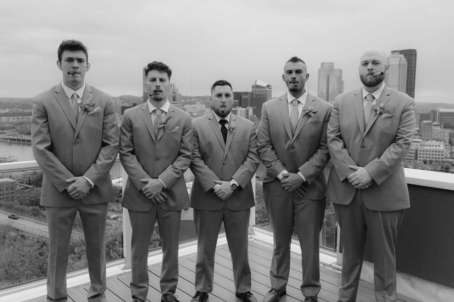 Five groomsmen in matching gray suits pose together on a rooftop with a city skyline backdrop.
