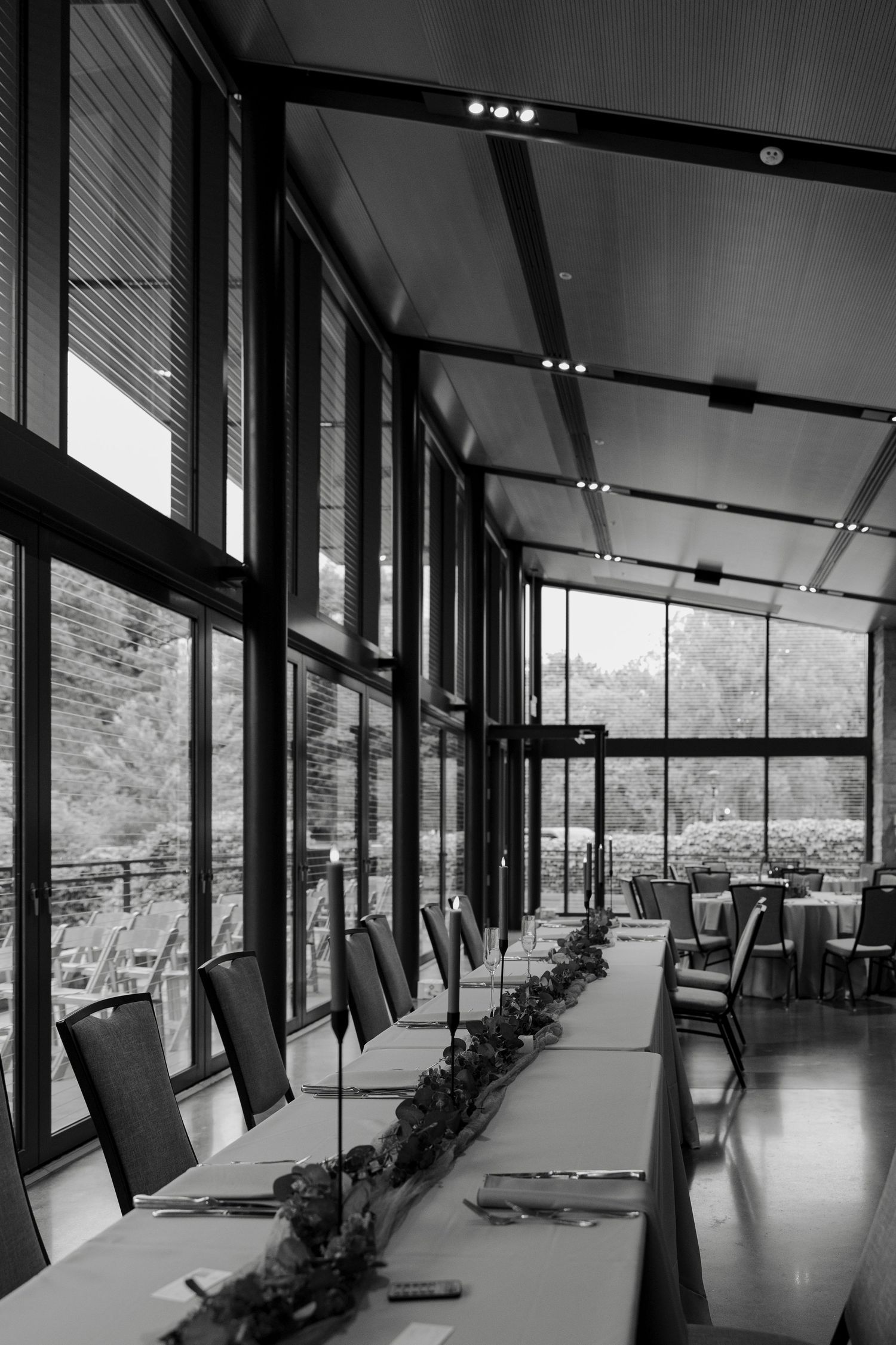 Black and white photo of an elegant dining room with floor-to-ceiling windows, long table, and chairs overlooking lush greenery.