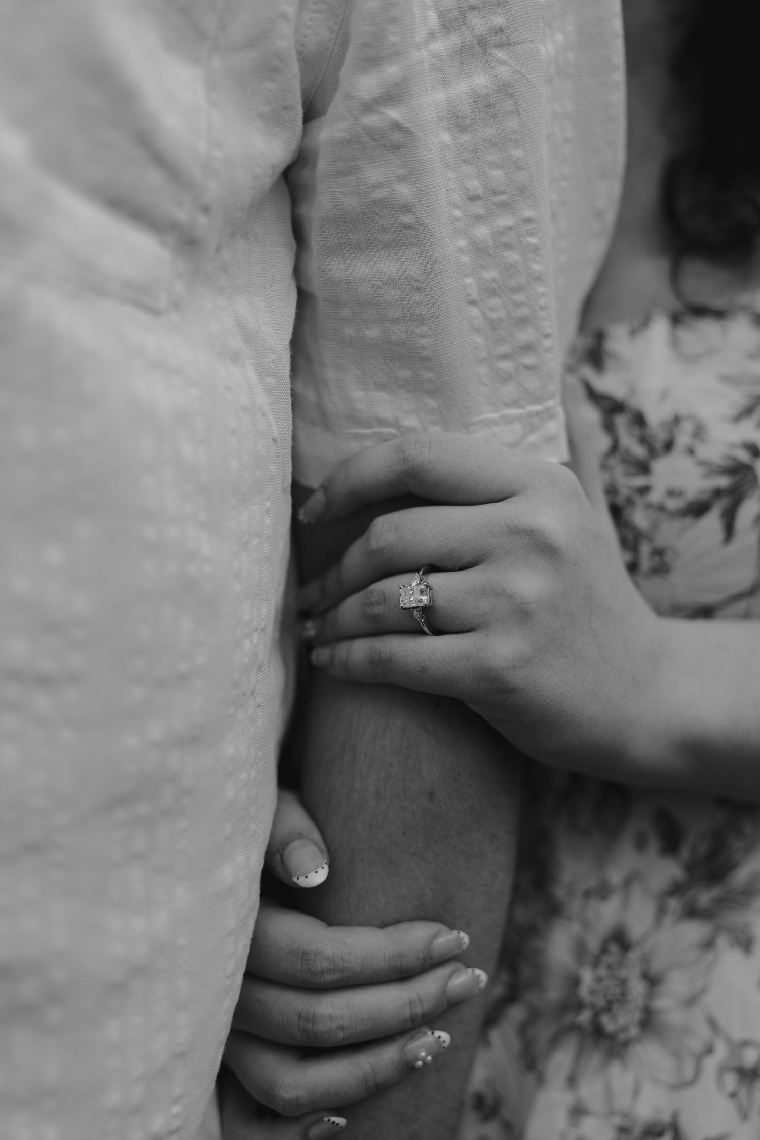 Close-up black and white photo of a woman's hand with a diamond engagement ring resting on a man's arm.