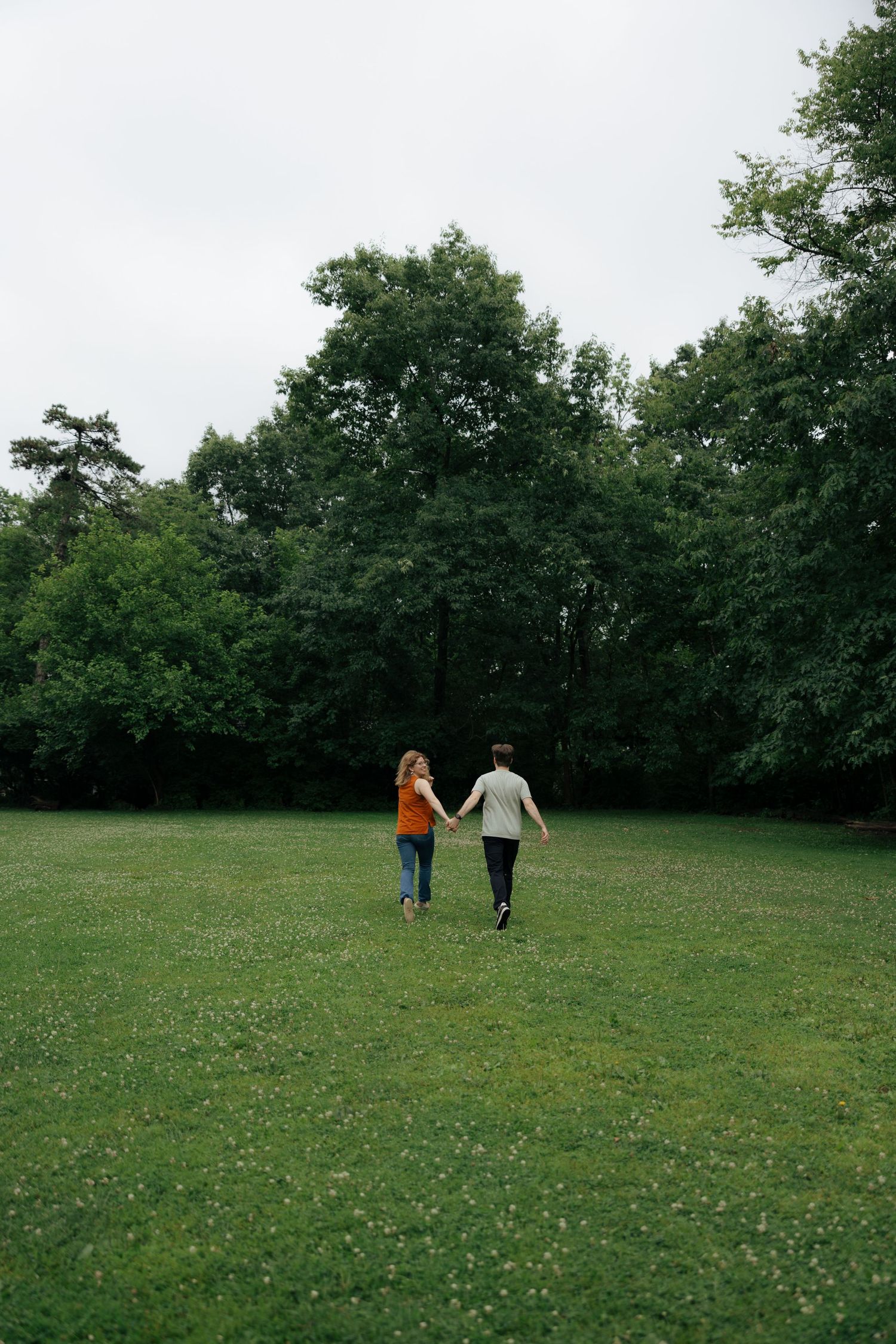 Couple holding hands walking through lush green park with tall trees on overcast day.