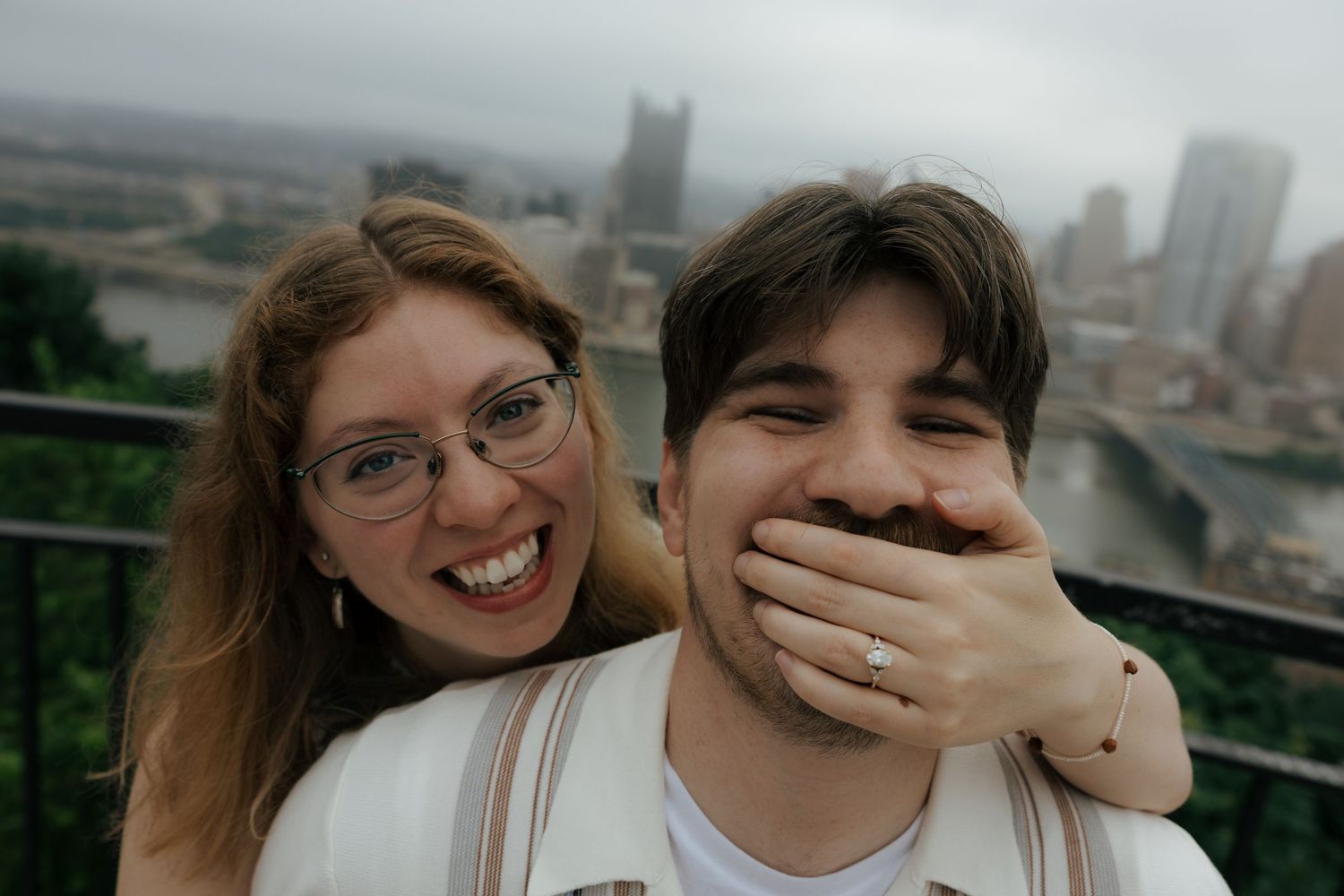 Joyful woman with glasses playfully covering her laughing partner's mouth with engagement ring visible.