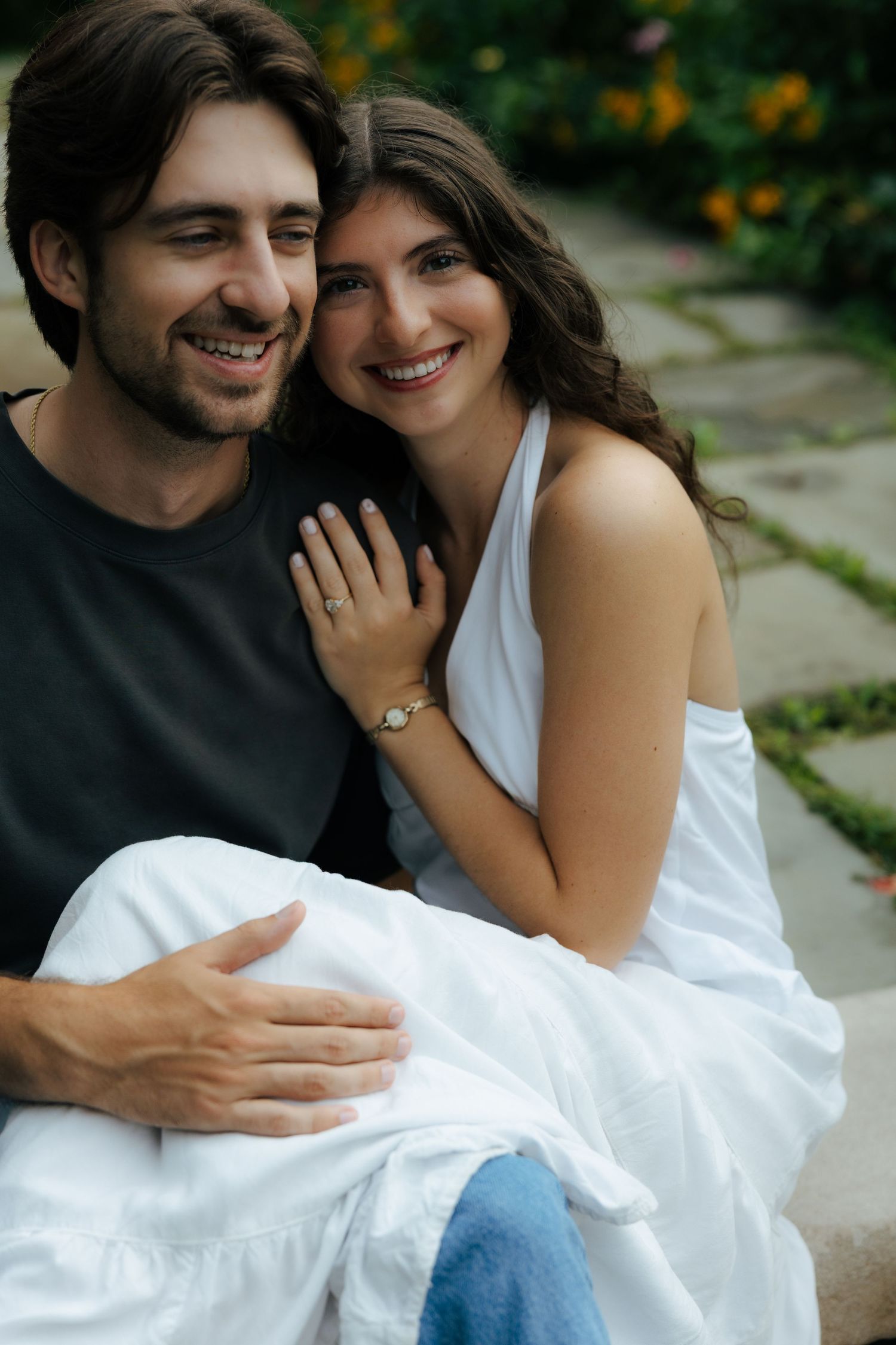 A smiling couple sits closely together outdoors surrounded by lush green garden foliage.