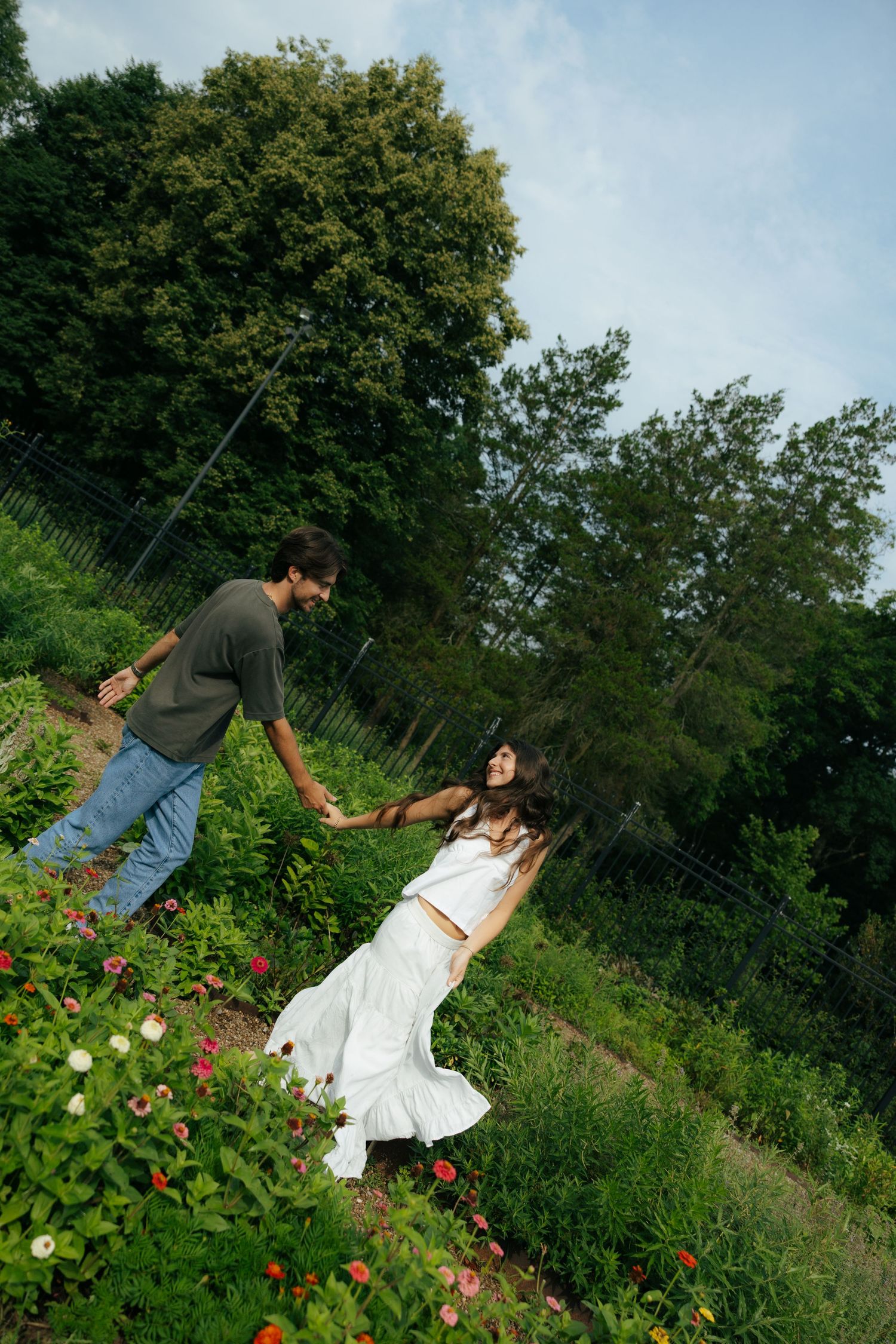 Couple holding hands and dancing playfully in a lush garden surrounded by colorful blooming flowers.