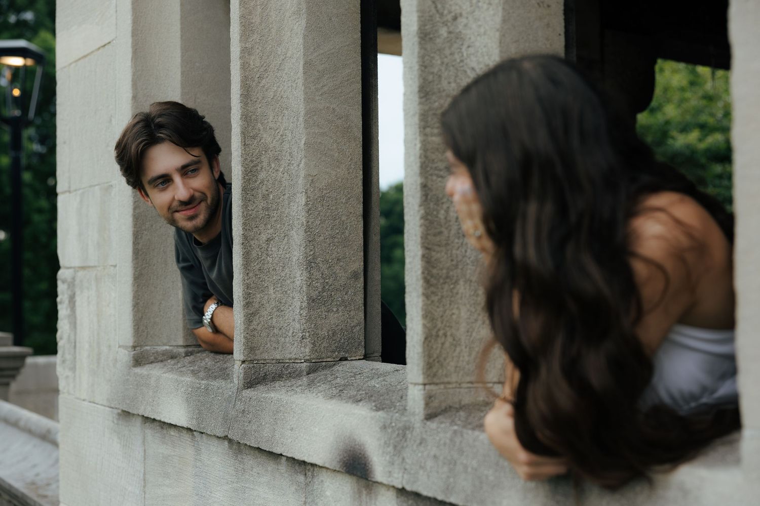 Smiling bearded man peering through stone columns at long-haired woman in white top at historic building.