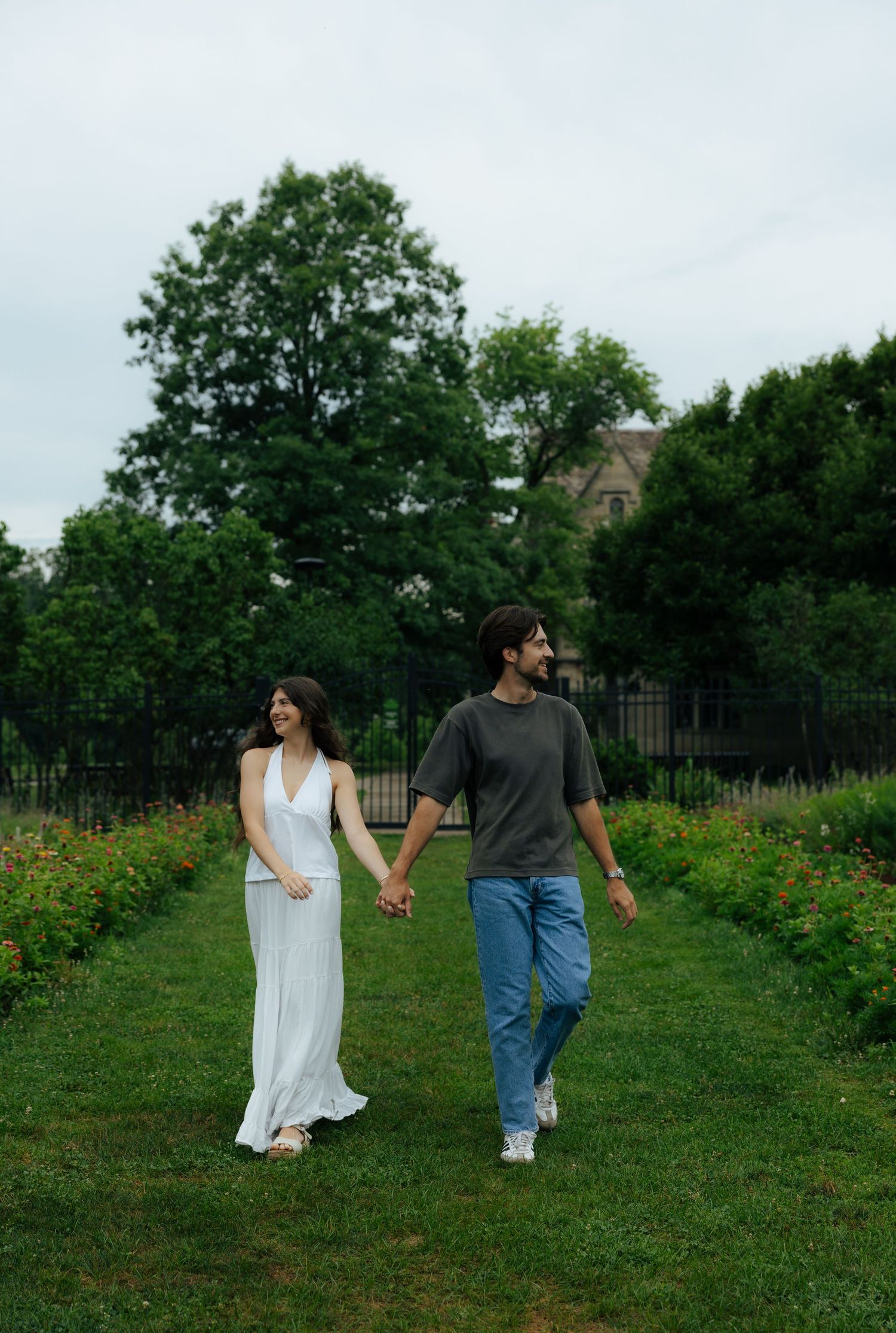 A couple holds hands and walks through a lush green garden path, woman in white dress, man in gray shirt and jeans.